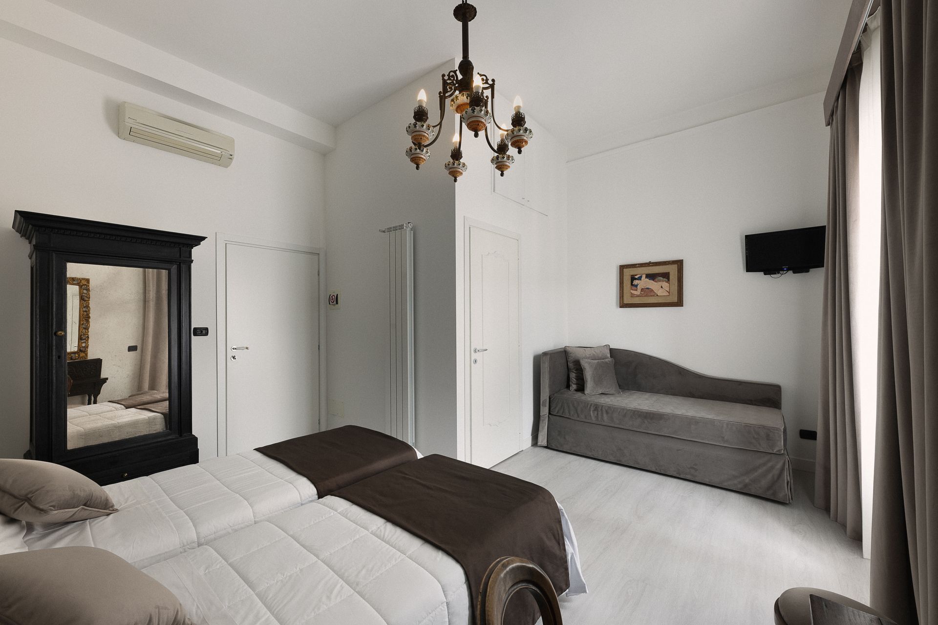 Bright bedroom with bed, gray sofa, mirror, chandelier, and large curtained window