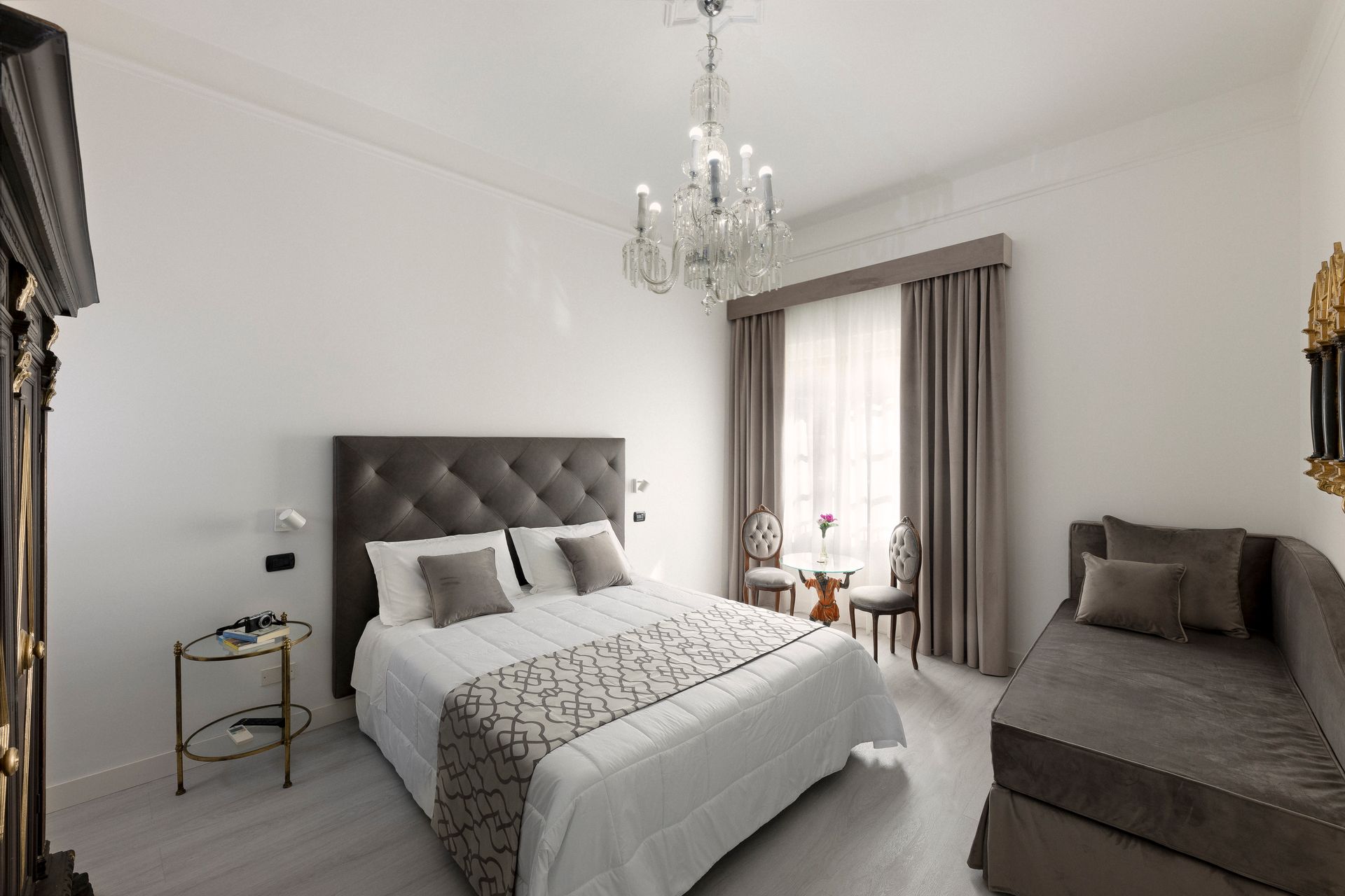 Bright bedroom with a tufted bed, chandelier, gray sofa, and window curtains