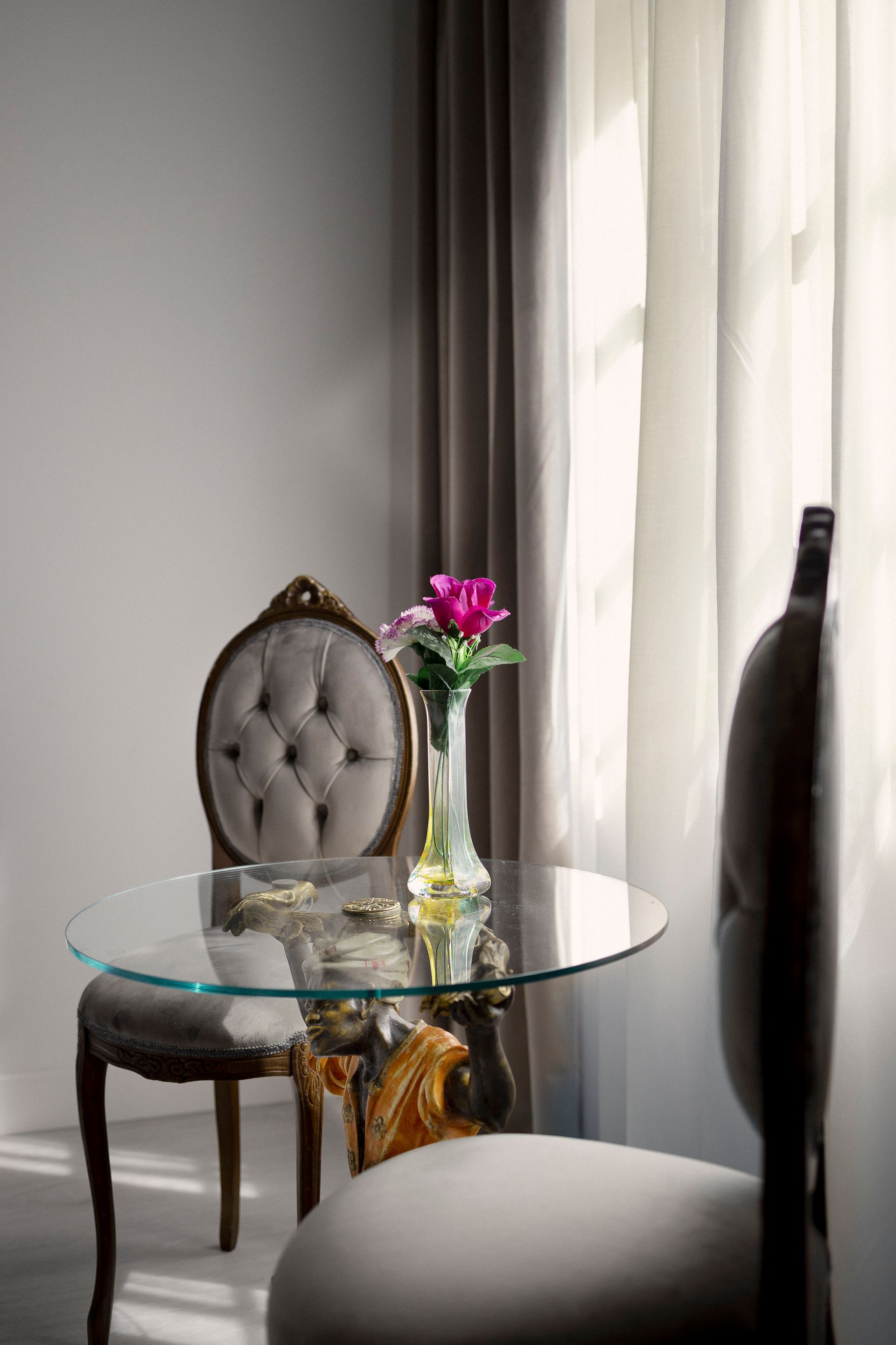 Elegante silla y mesa de cristal junto a una ventana, con un jarrón de flores rosas y una botella oscura.