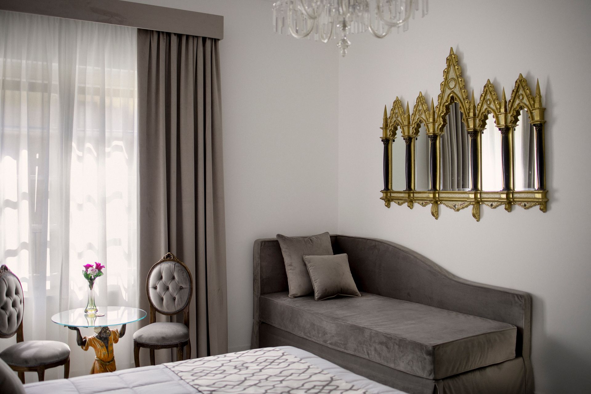 Elegant bedroom with gray chaise lounge, gold wall mirrors, sheer curtains, and a small round table by the window