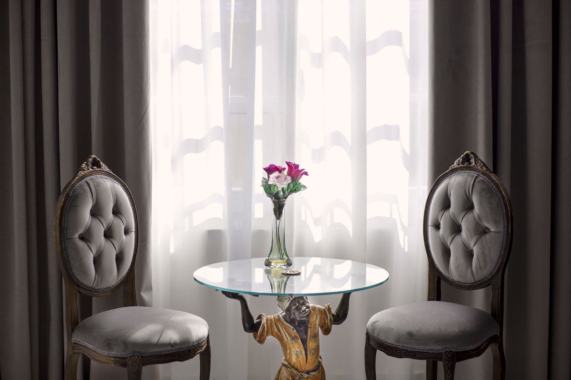 Two gray tufted chairs flank a glass table with a vase of pink flowers by sheer curtains.