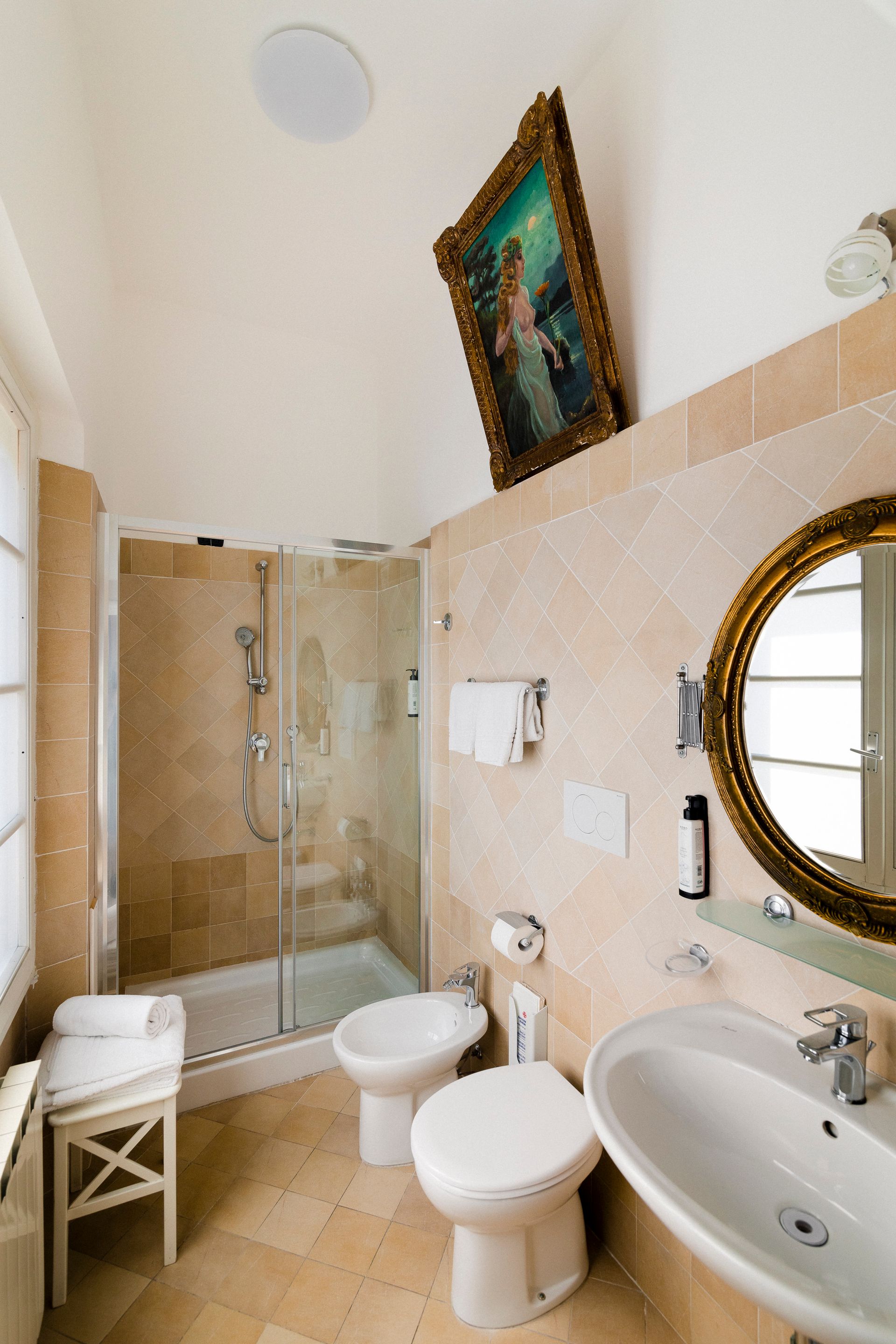 Small bathroom with glass shower, bidet, toilet, sink, and ornate mirror under a high ceiling.