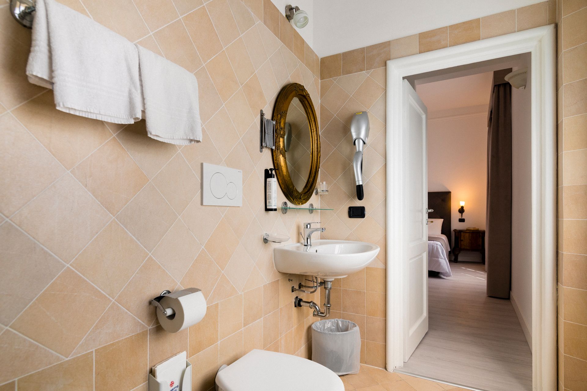 Compact beige bathroom with wall-mounted sink, towel rack, mirror, and open door to a bedroom.
