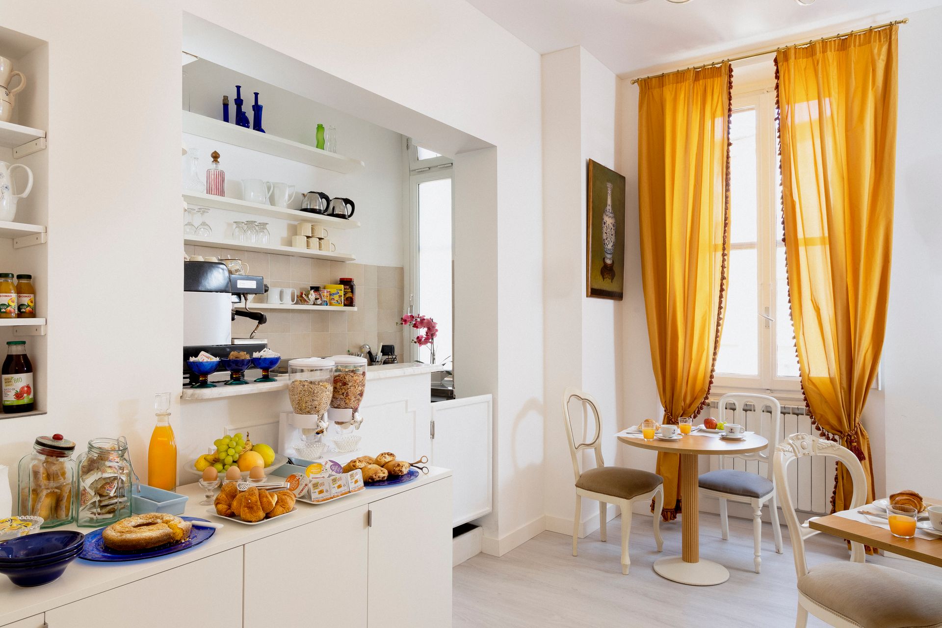 Bright white kitchen-dining nook with orange curtains, open shelves, and a small table by the window
