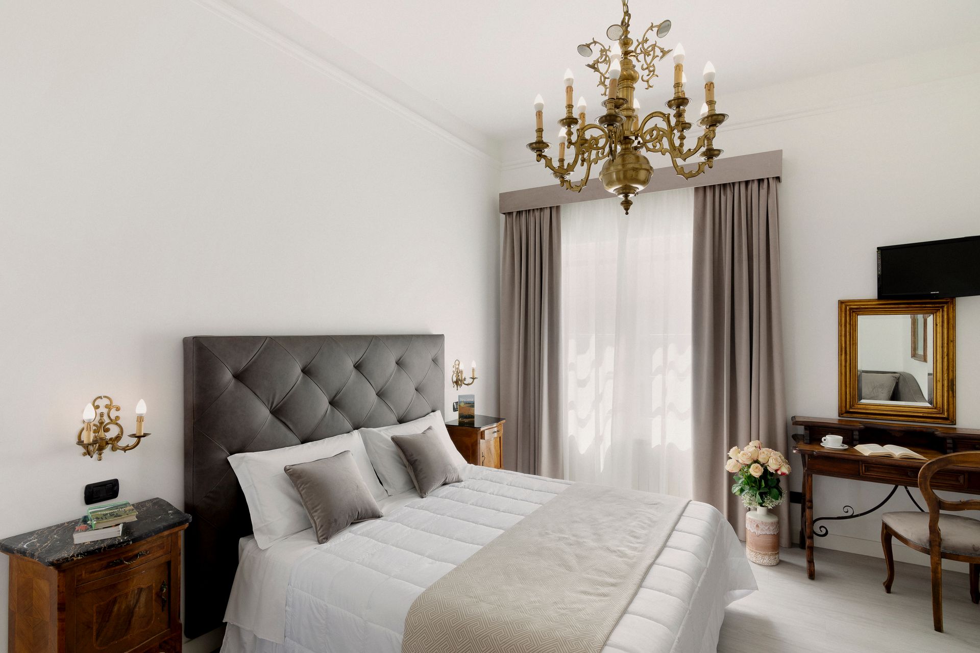Elegant bedroom with tufted gray bed, gold chandelier, sheer curtains, and antique desk by window