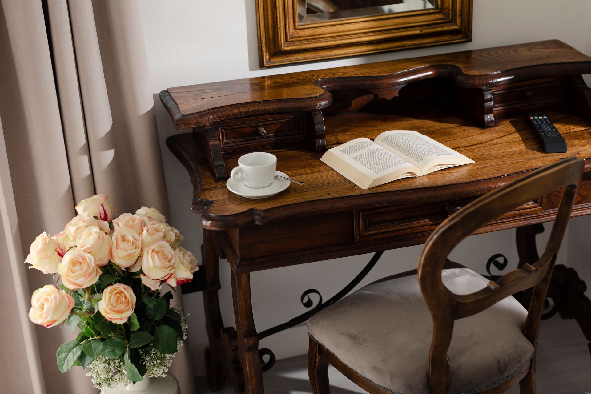 Elegant wooden writing desk with open book and teacup beside a vase of pale roses in a room