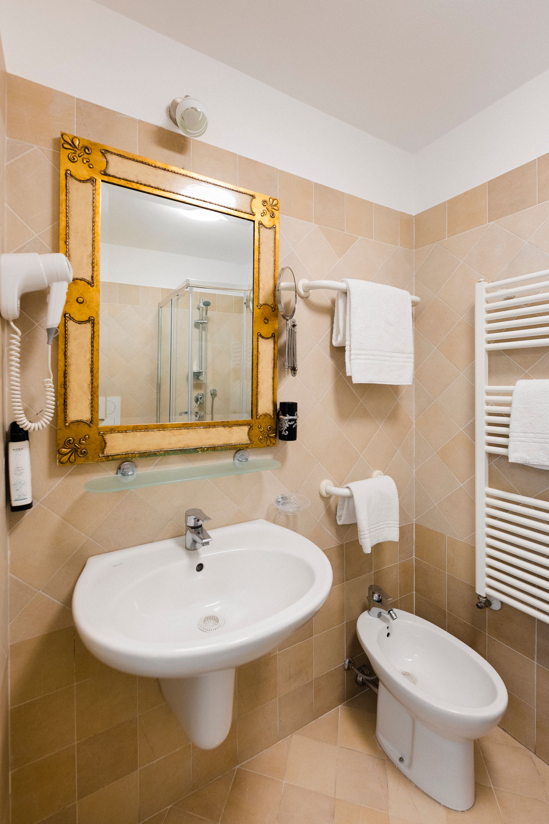 Small beige bathroom with sink, bidet, towel rack, and ornate gold-framed mirror