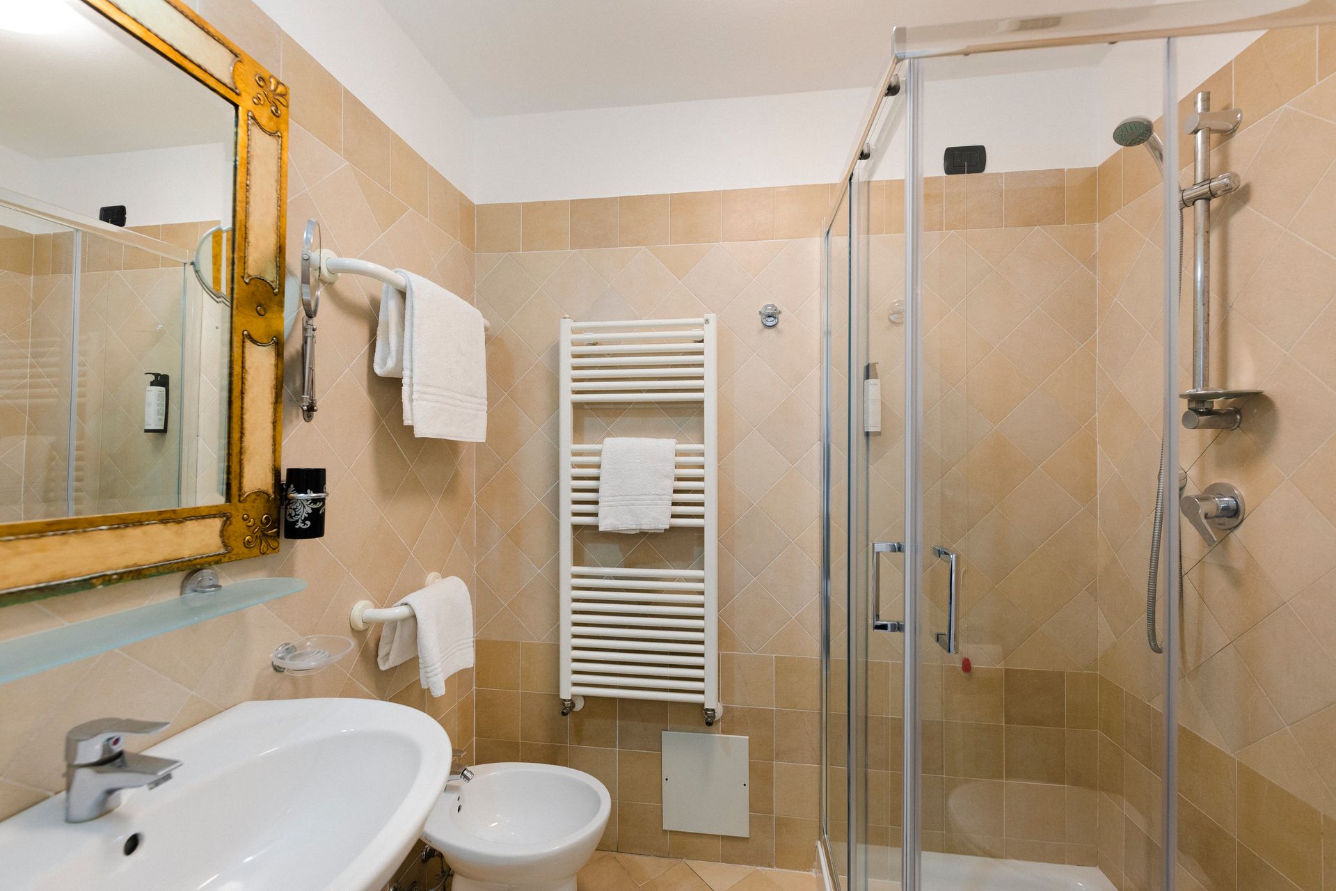 Compact beige bathroom with sink, mirror, towel rack, toilet, and glass shower enclosure