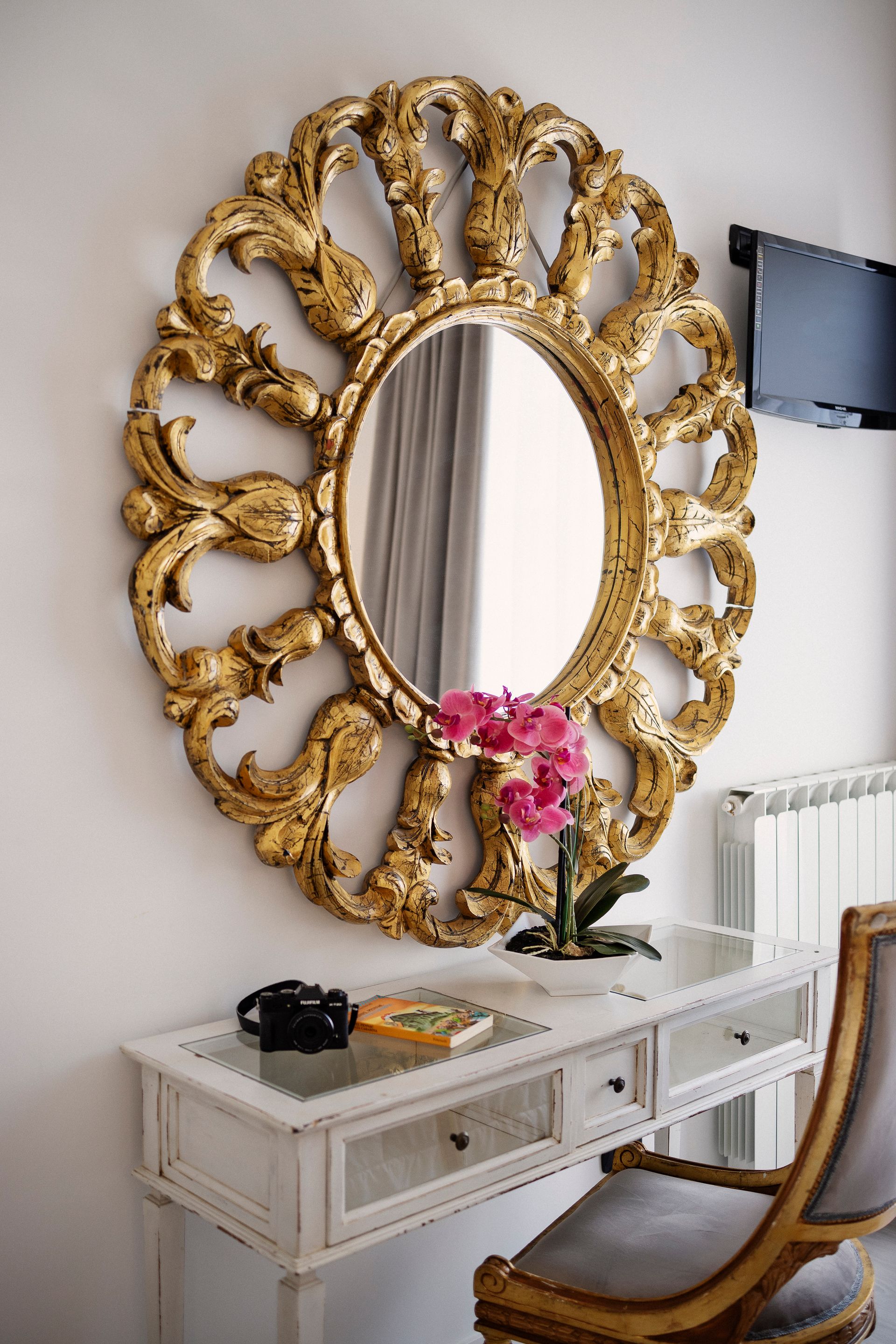 Ornate gold-framed mirror above a white console table with pink flowers in a bright room
