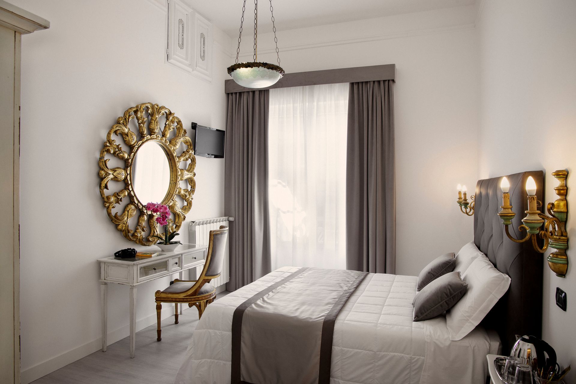 Bright bedroom with a bed, vanity, gold mirror, and gray curtains by a window.