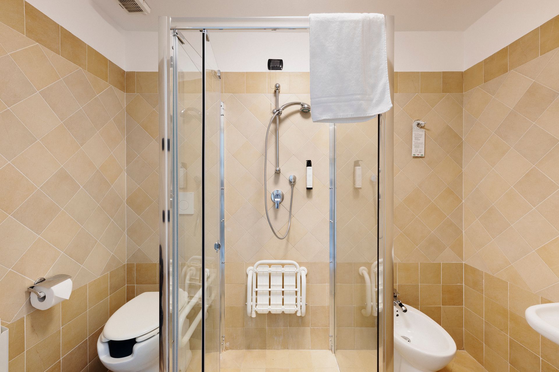 Accessible bathroom with glass shower, wall-mounted toilet, and beige tile walls