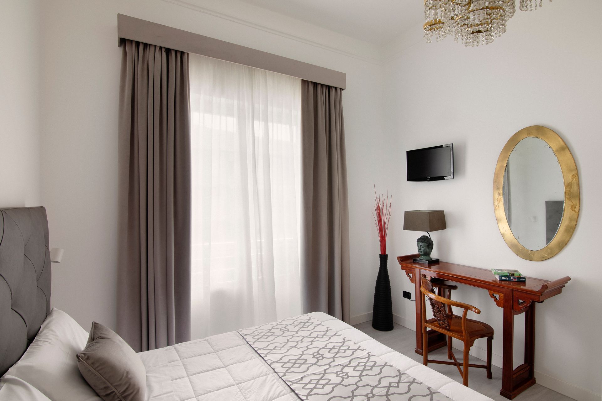 Bedroom with gray bed, sheer curtains, small wooden desk, round gold mirror, and wall-mounted TV
