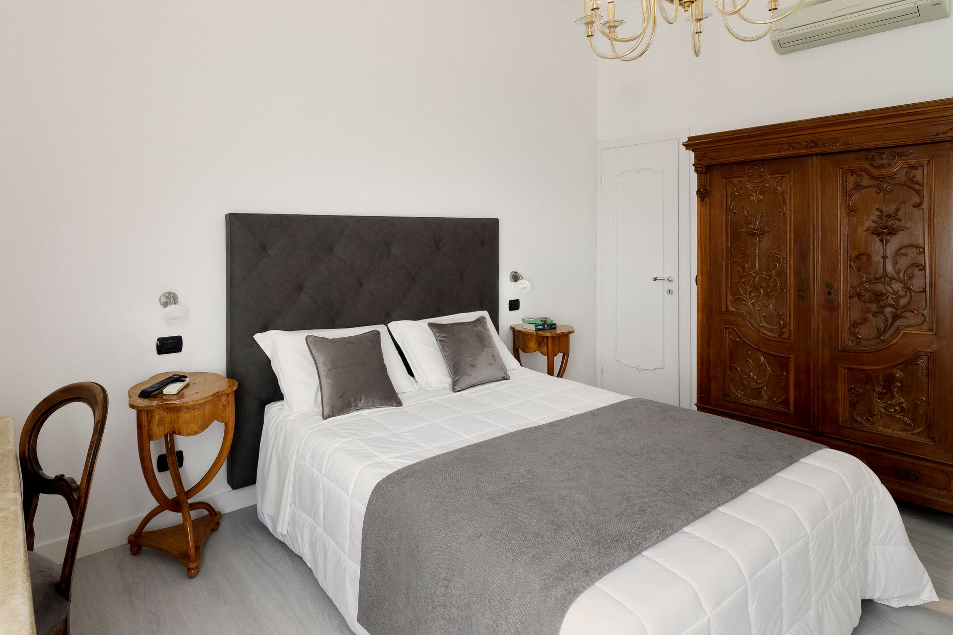 Bright bedroom with a gray bed, wooden armoire, and small side tables against white walls.