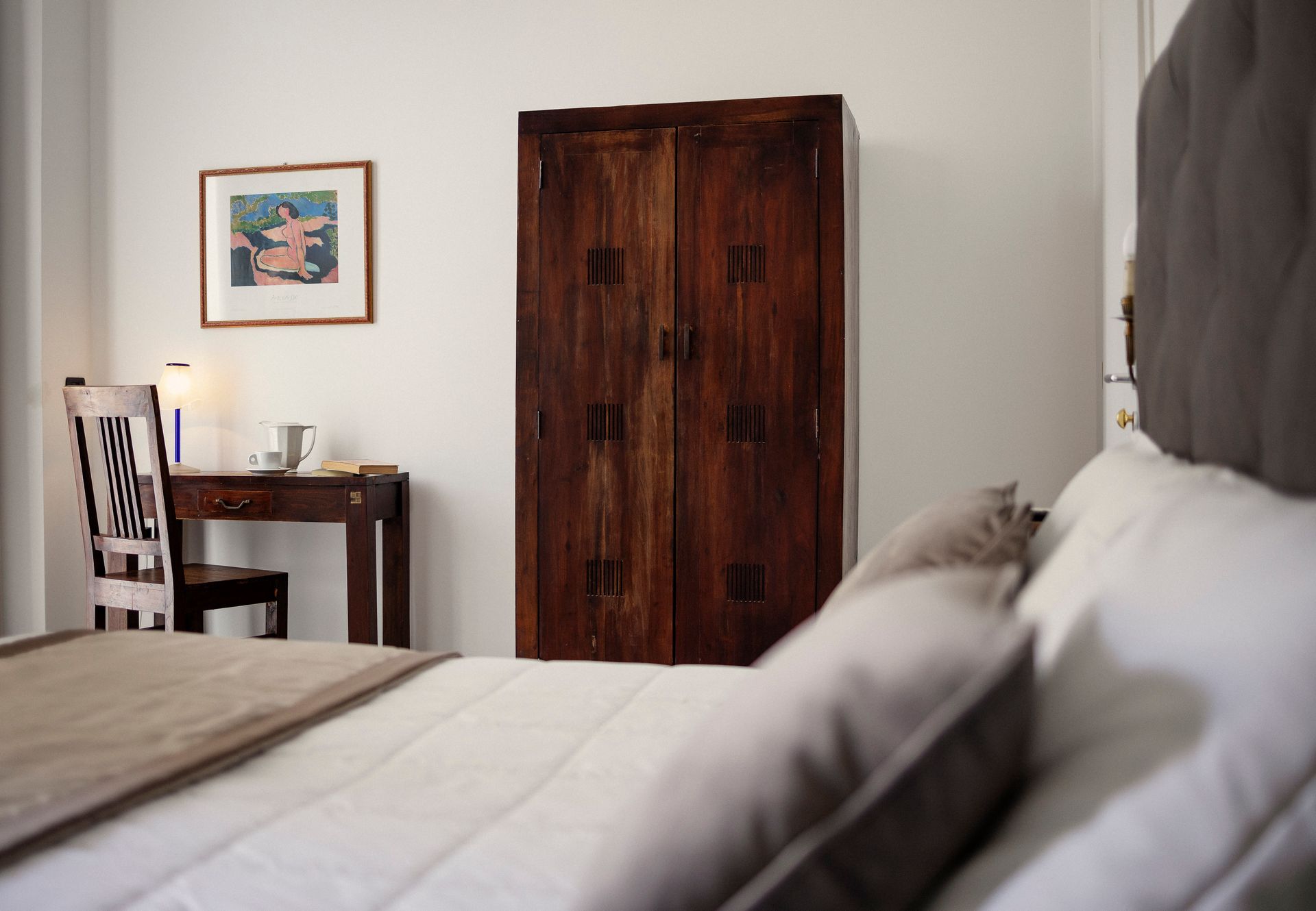 Bedroom with a wooden wardrobe, desk, chair, framed art, and a bed in the foreground.