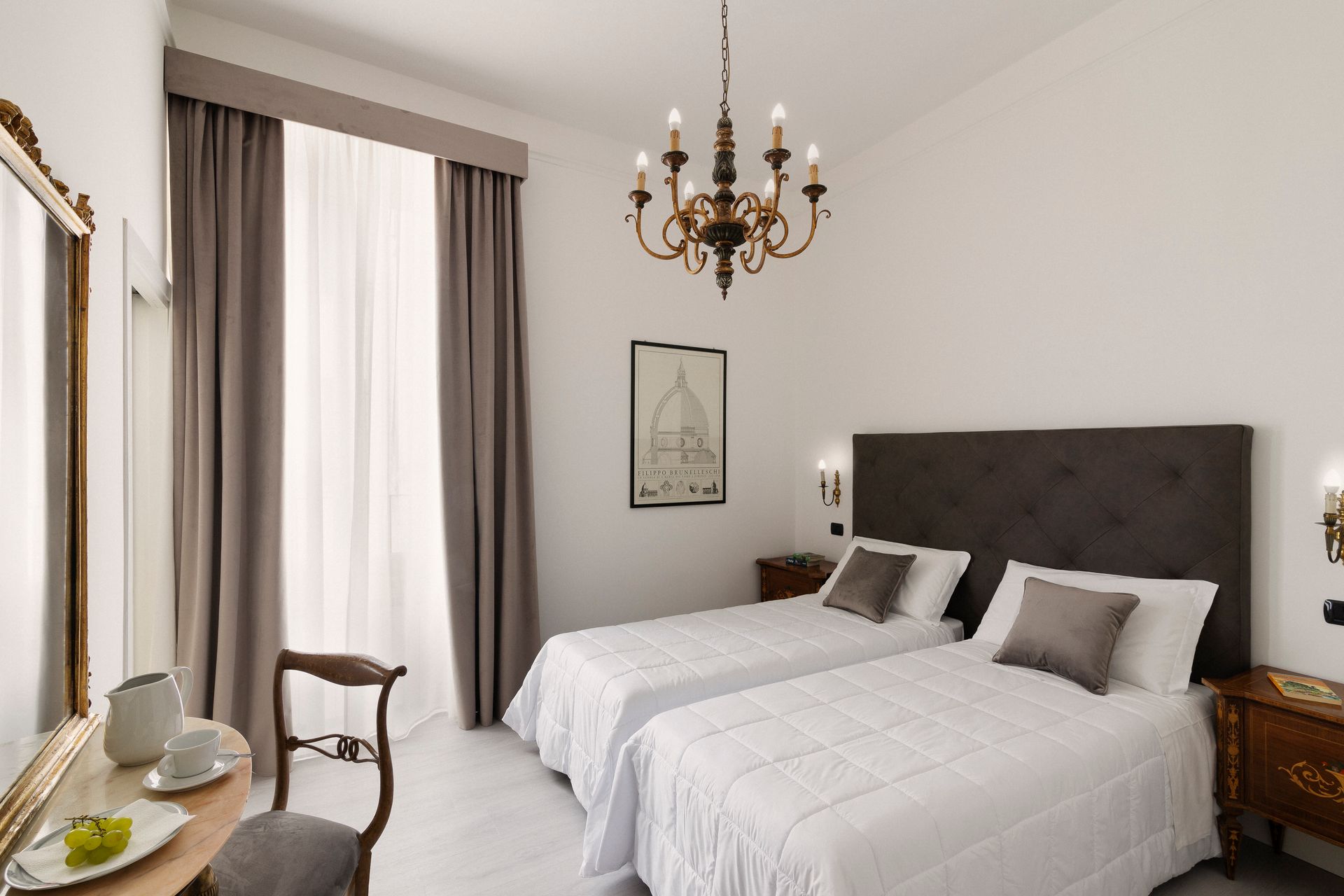 Bright bedroom with two single beds, chandelier, and a desk by a window with curtains.