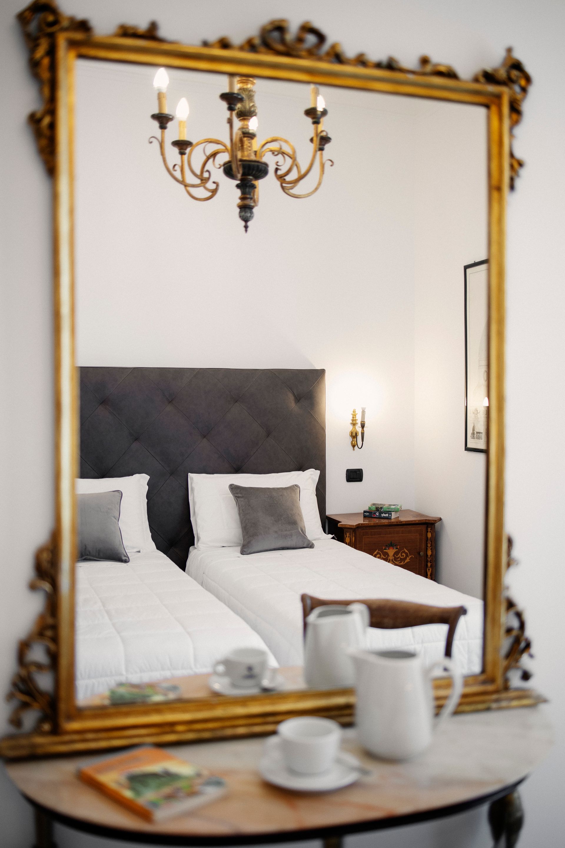 Elegant bedroom seen in a gold-framed mirror, with a bed, bedside lamp, and tray on the table.