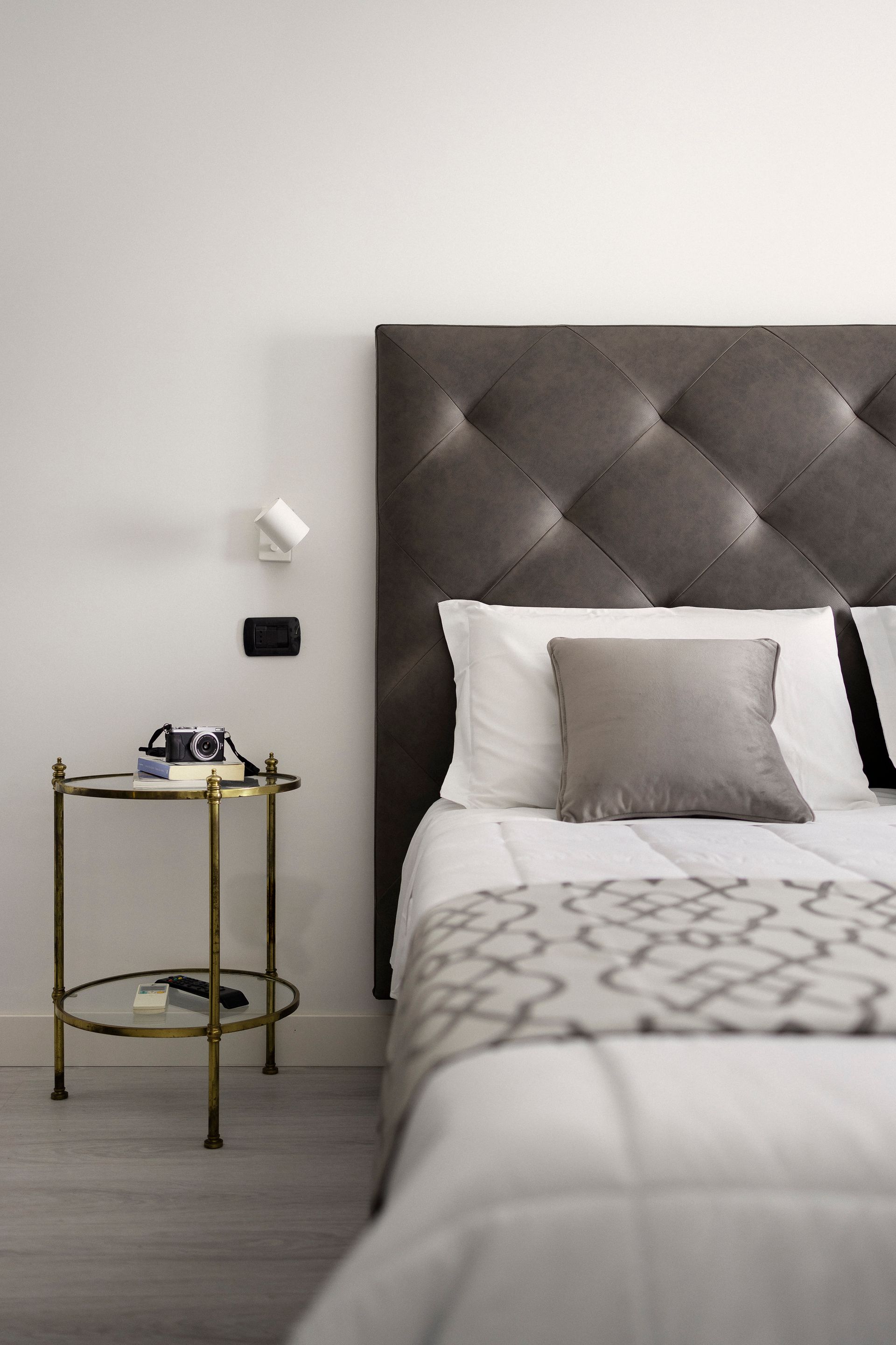 Dormitorio moderno con cabecero gris capitoné, ropa de cama blanca y una pequeña mesita de noche dorada.