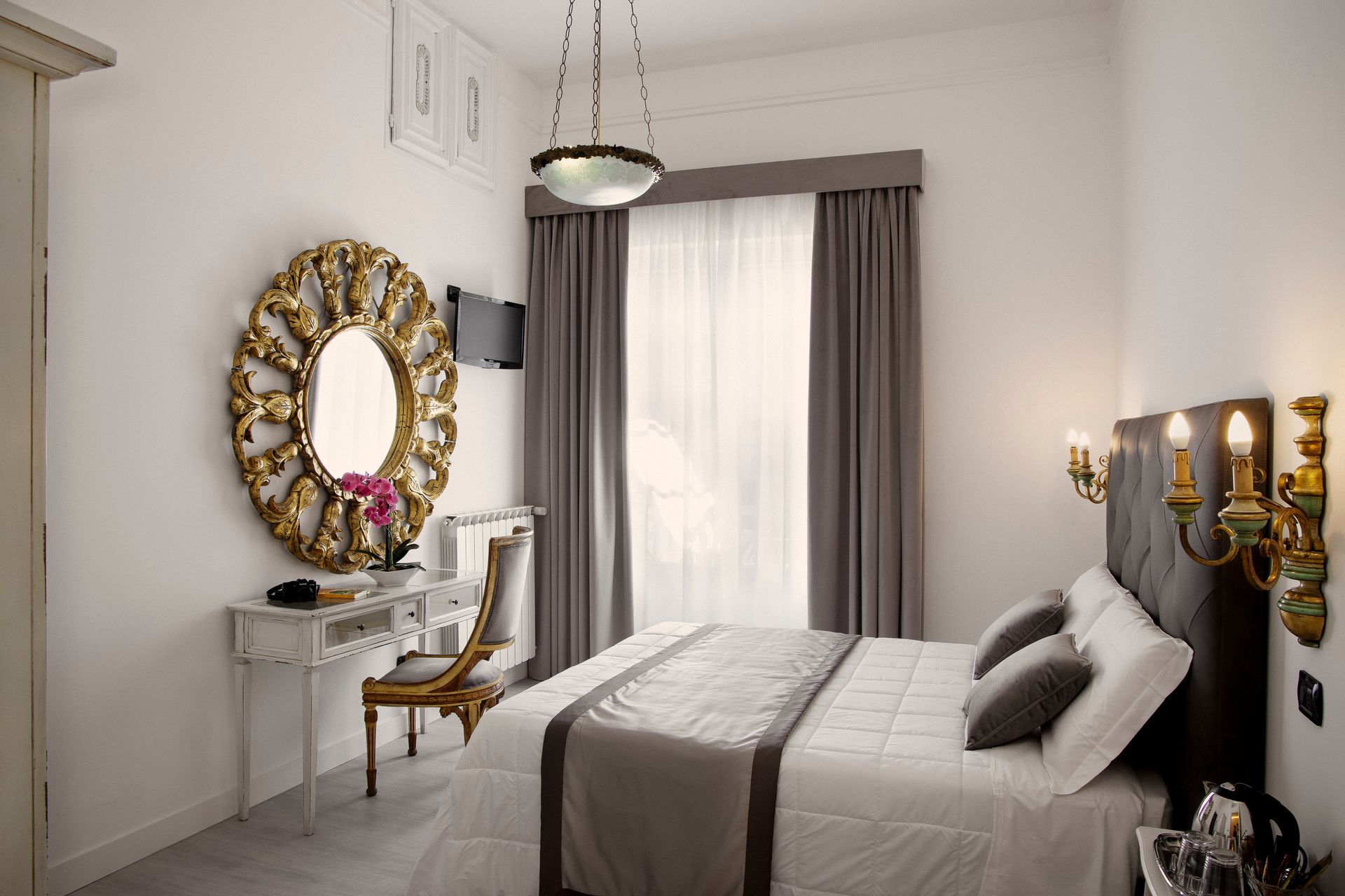 Habitación luminosa con cama, escritorio y silla blancos, espejo grande dorado y cortinas grises.