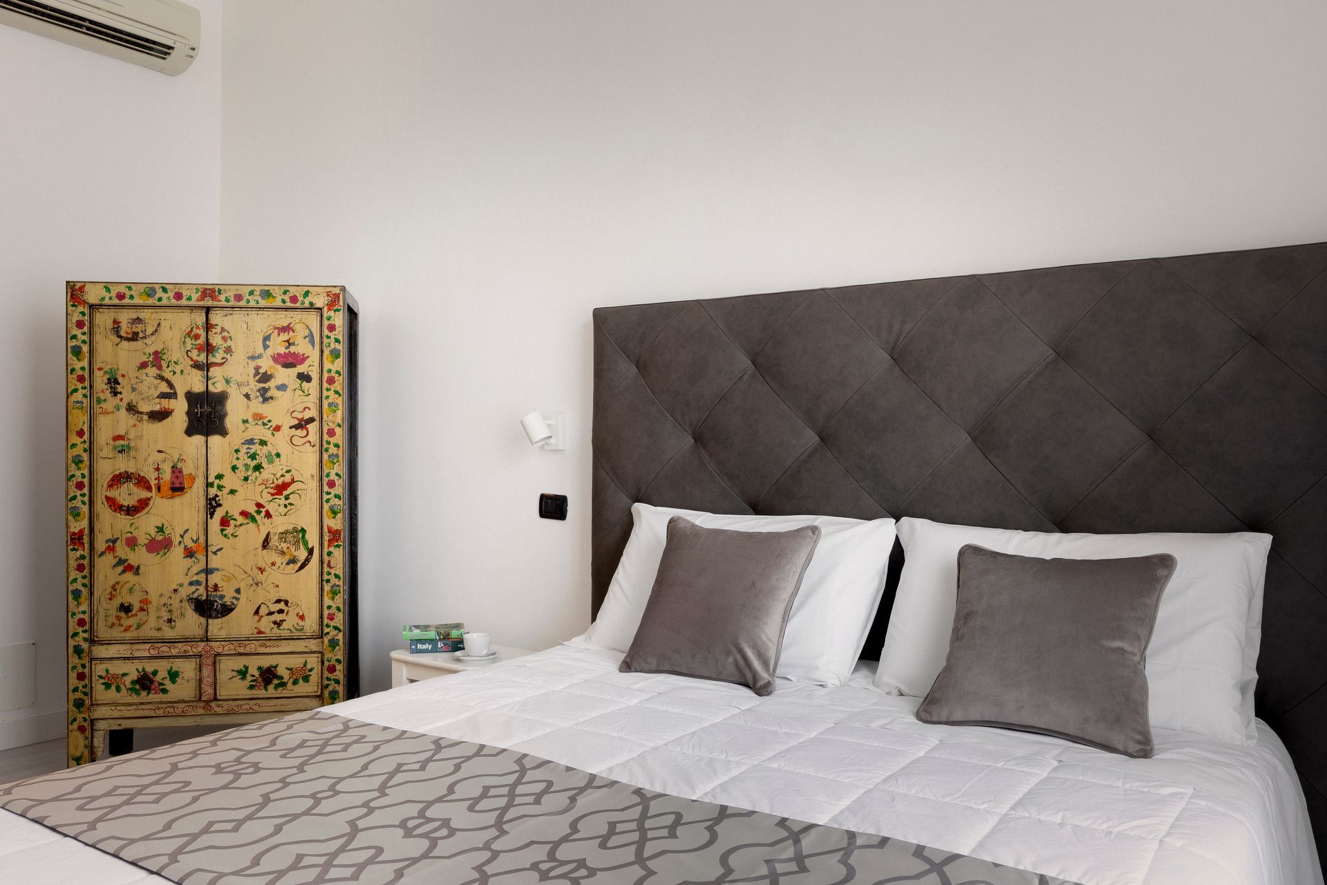 Habitación luminosa con cabecero acolchado gris, ropa de cama blanca y armario con estampado floral colorido.