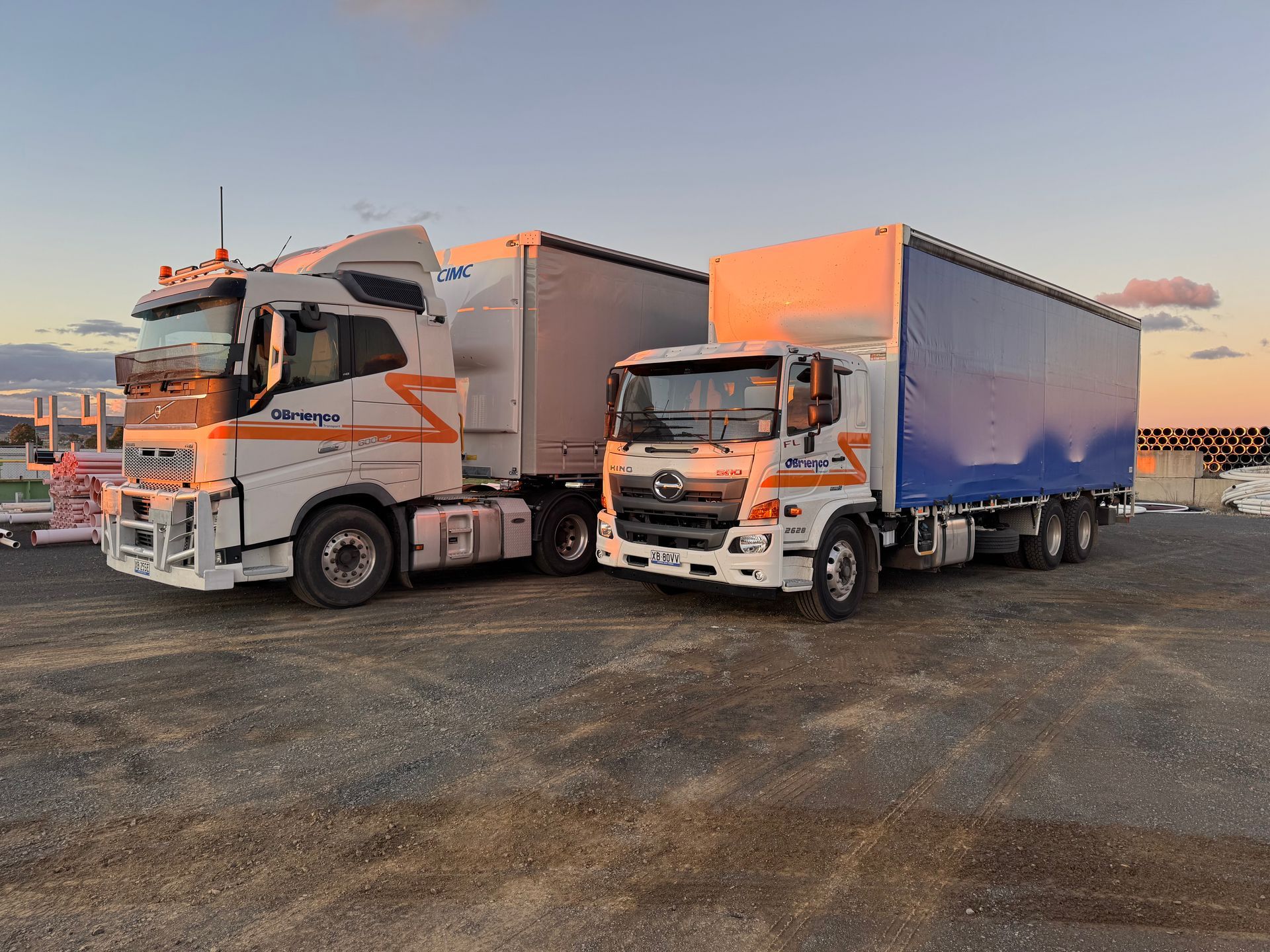 Two white trucks parked on gravel at sunset; one larger with white trailer, one smaller with blue.