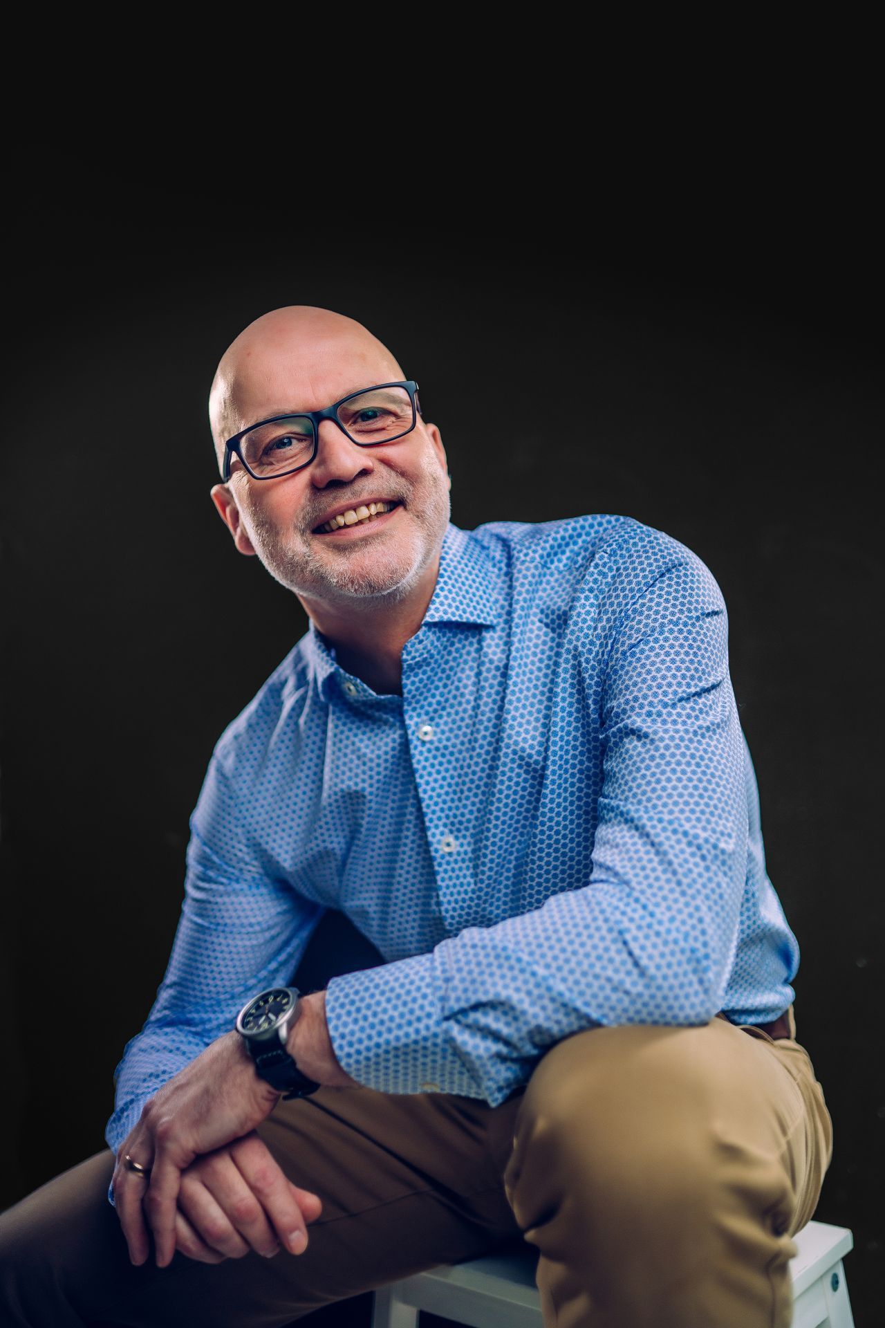 A man wearing glasses and a blue shirt is sitting on a stool.