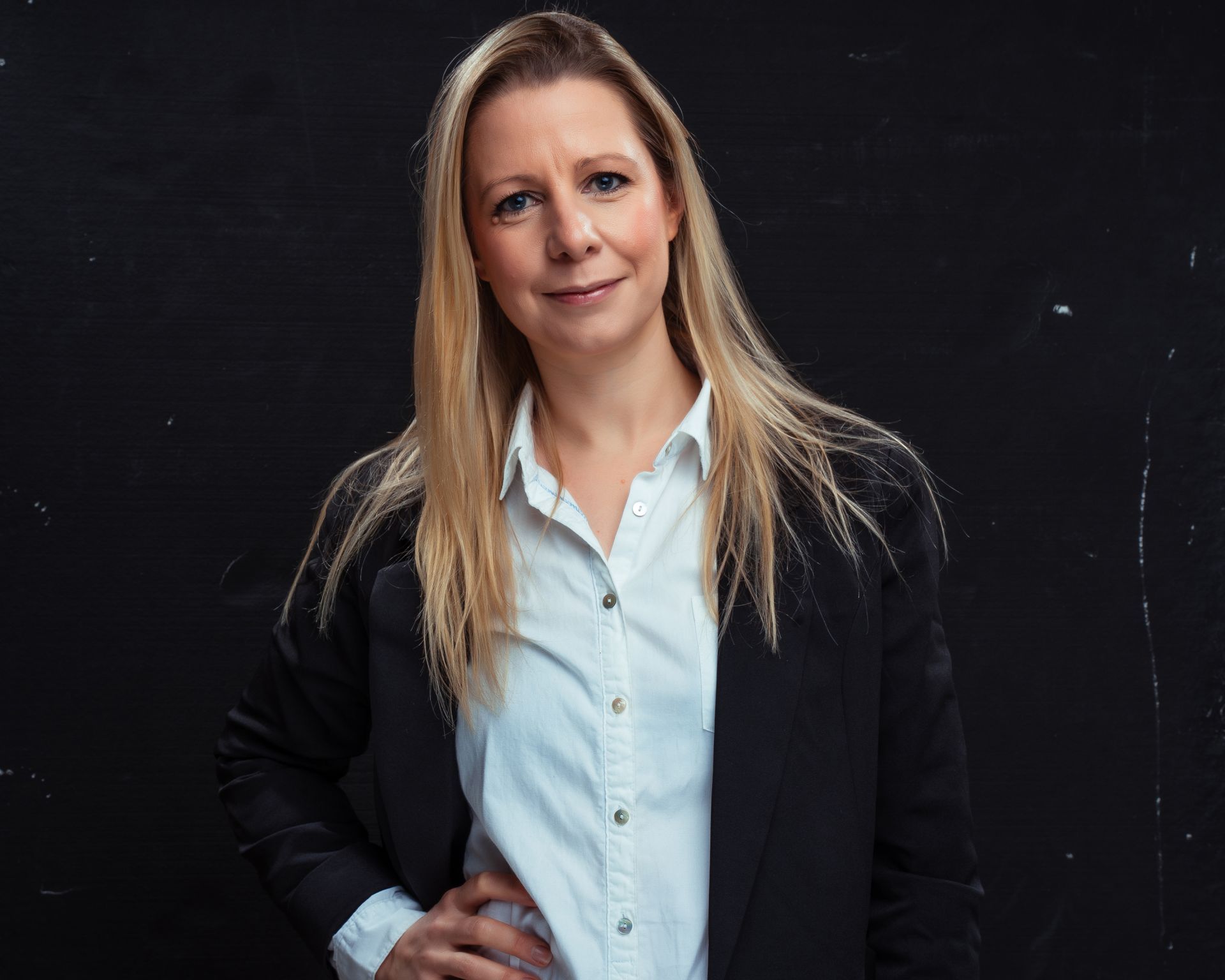 A woman in a black jacket and white shirt is standing in front of a black wall.