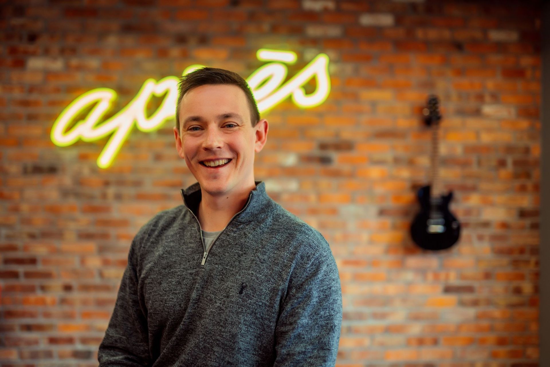 A man is smiling in front of a brick wall and a neon sign.