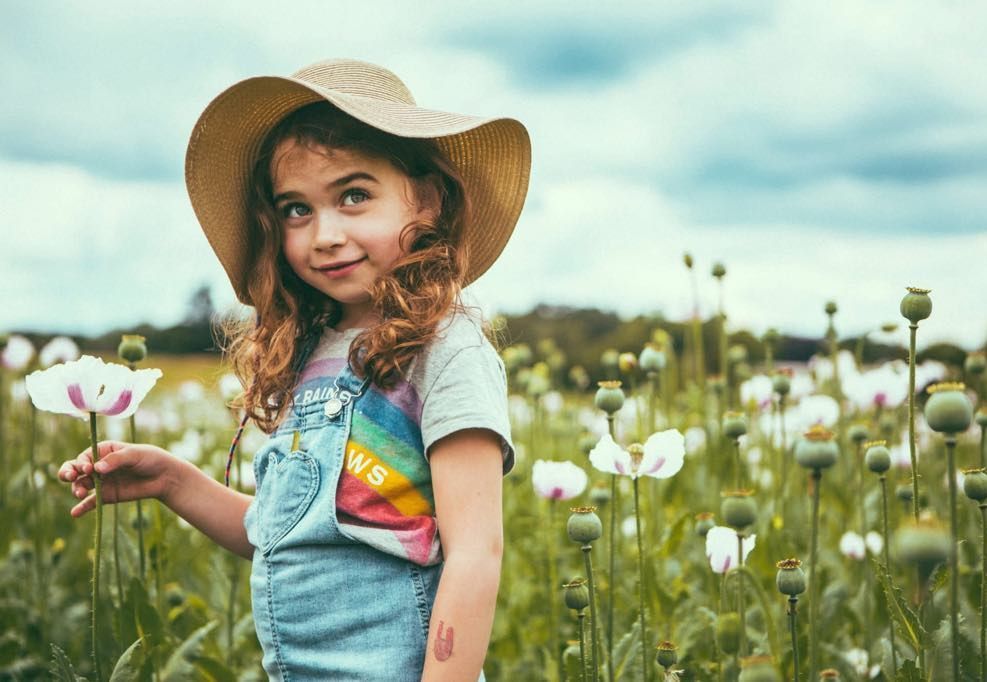 A little girl wearing a hat and overalls is standing in a field of flowers.
