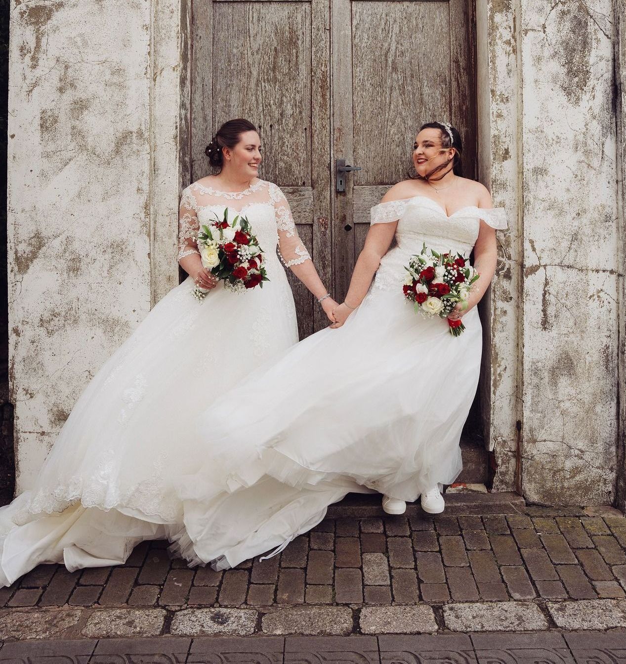 Two women in wedding dresses holding hands in front of a door