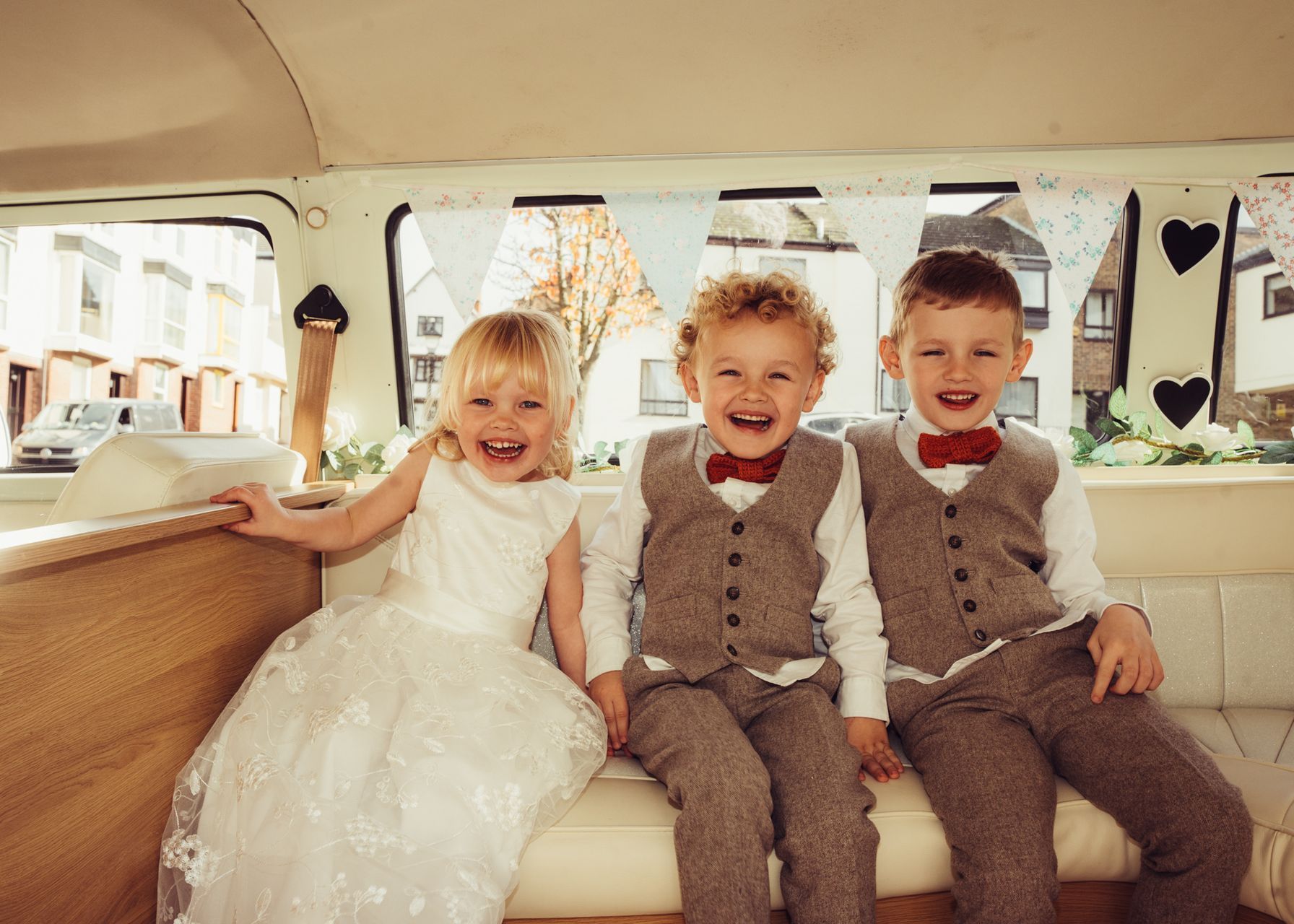 A flower girl and two ring bearers are sitting in the back of a wedding car.