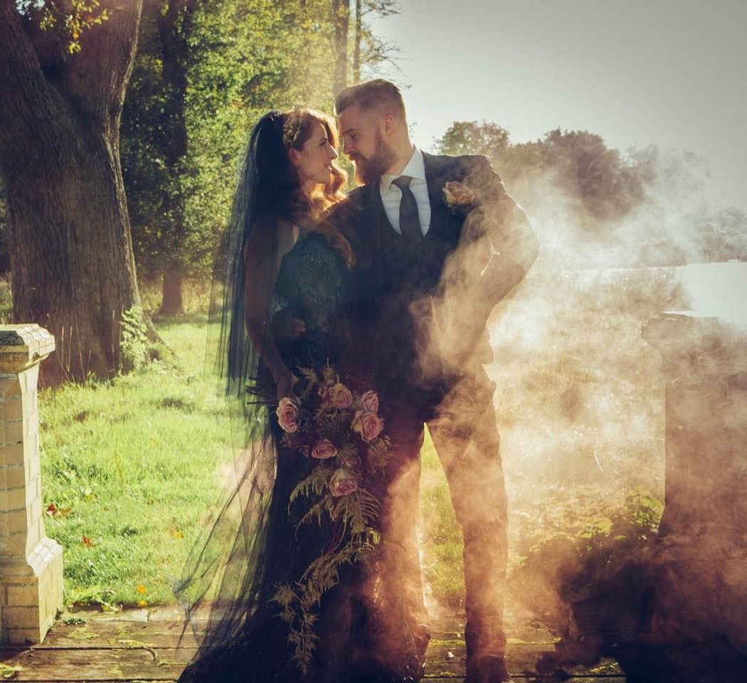 A bride and groom are standing next to each other in a field surrounded by smoke.