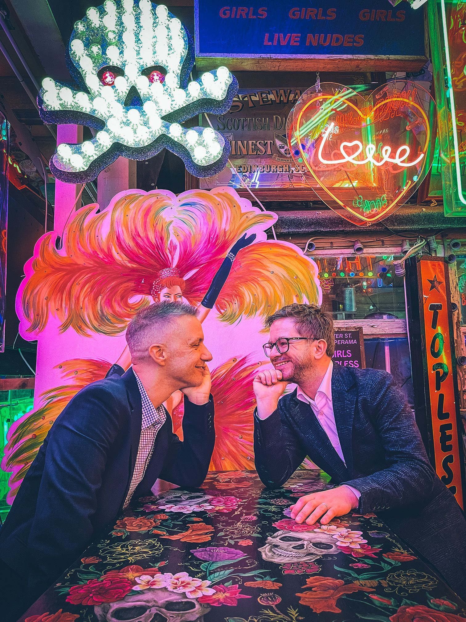 Two men are sitting at a table in front of a neon sign that says `` love ''.