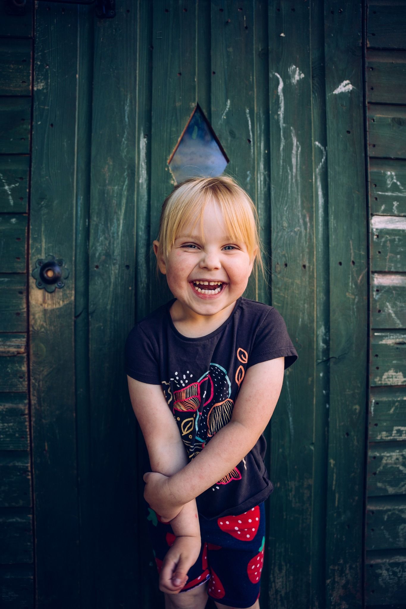 A little girl is standing in front of a green door and smiling.