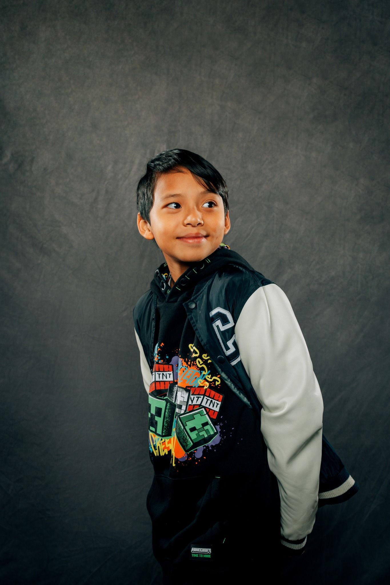 A young boy wearing a black and white hoodie is standing in front of a gray background.