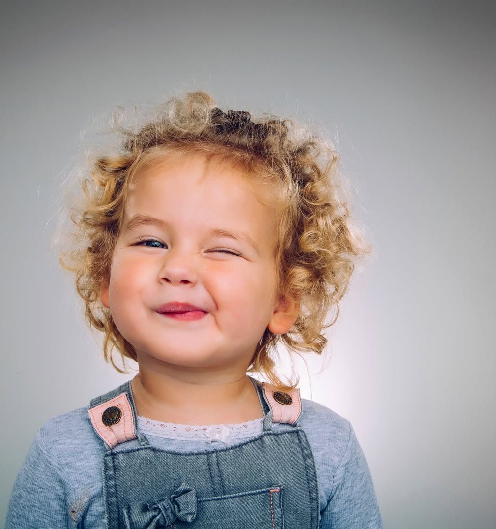 A little girl with curly hair is wearing overalls and smiling.