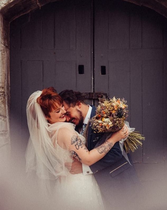 A bride and groom are hugging each other in front of a door while holding a bouquet of flowers.