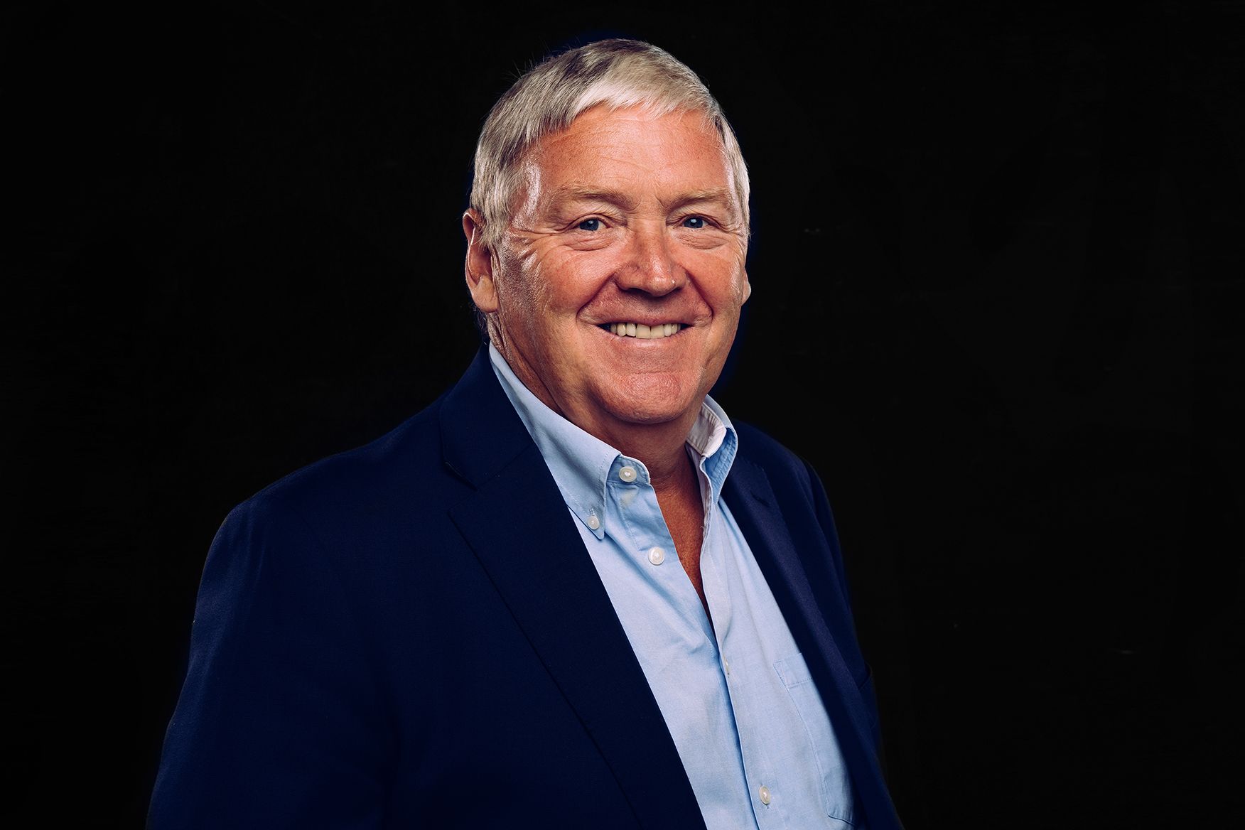 A man in a suit and blue shirt is smiling in front of a black background.
