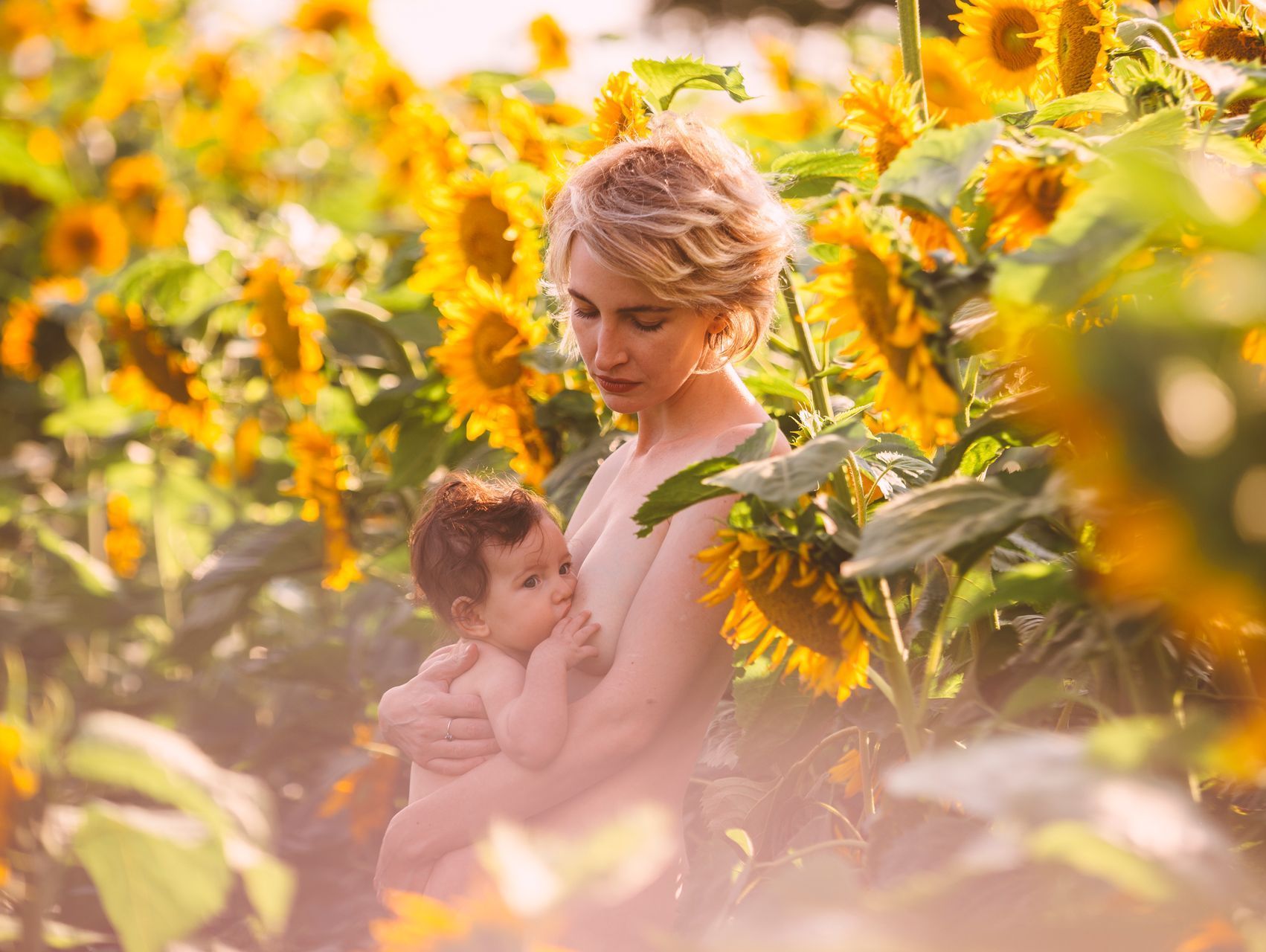 A woman is breastfeeding her baby in a field of sunflowers.