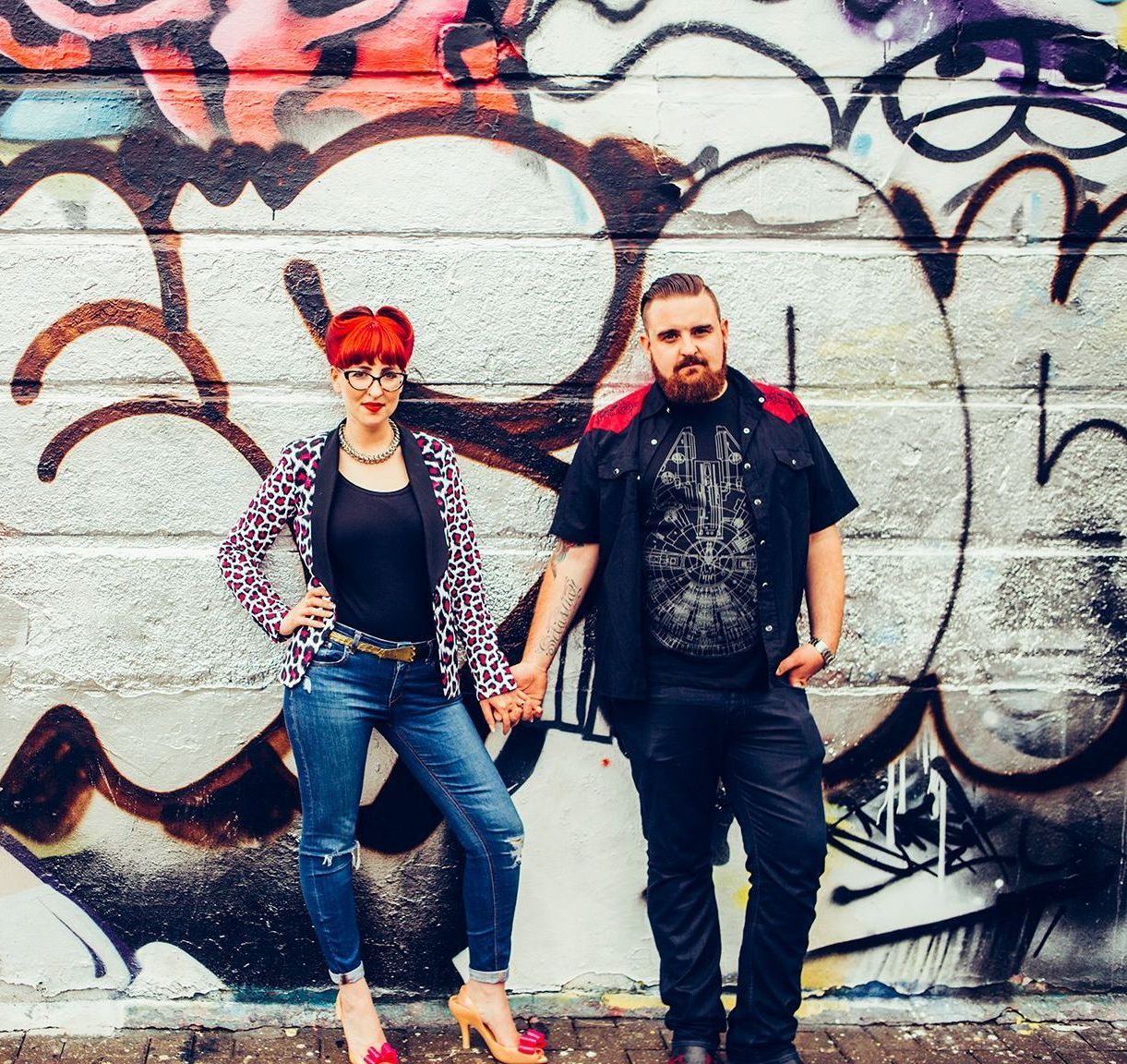 A man and a woman are standing in front of a wall with graffiti on it.