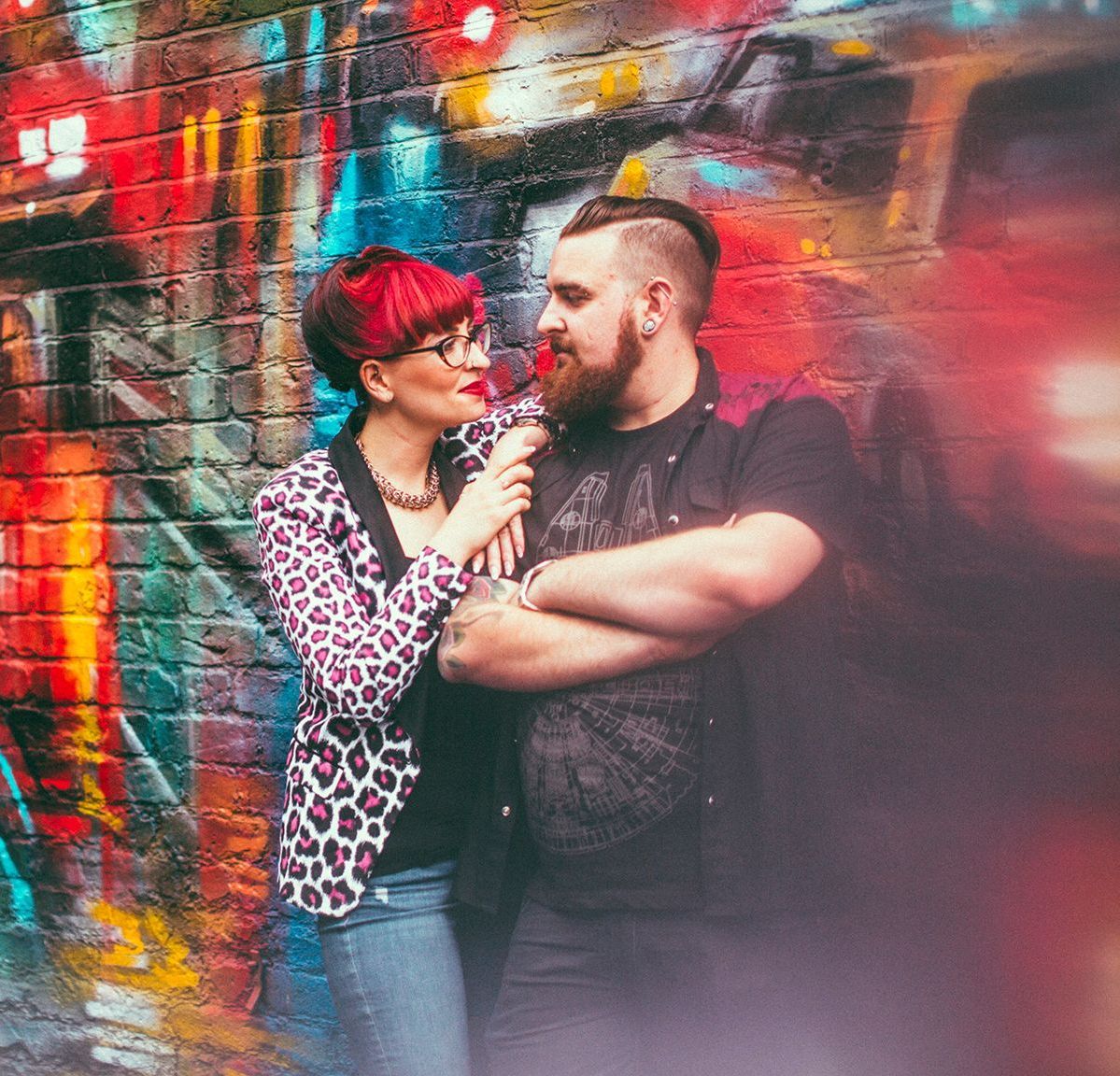 A man and a woman are standing in front of a brick wall with graffiti on it.