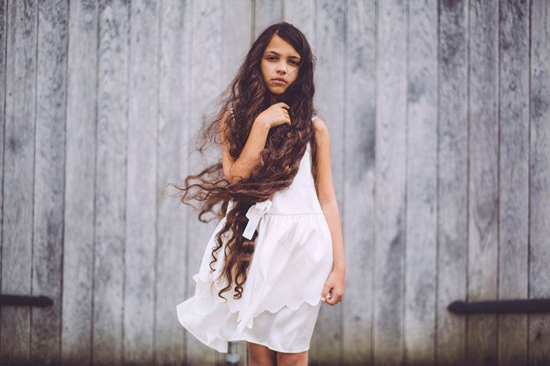 A young girl in a white dress with long hair is standing in front of a wooden wall.