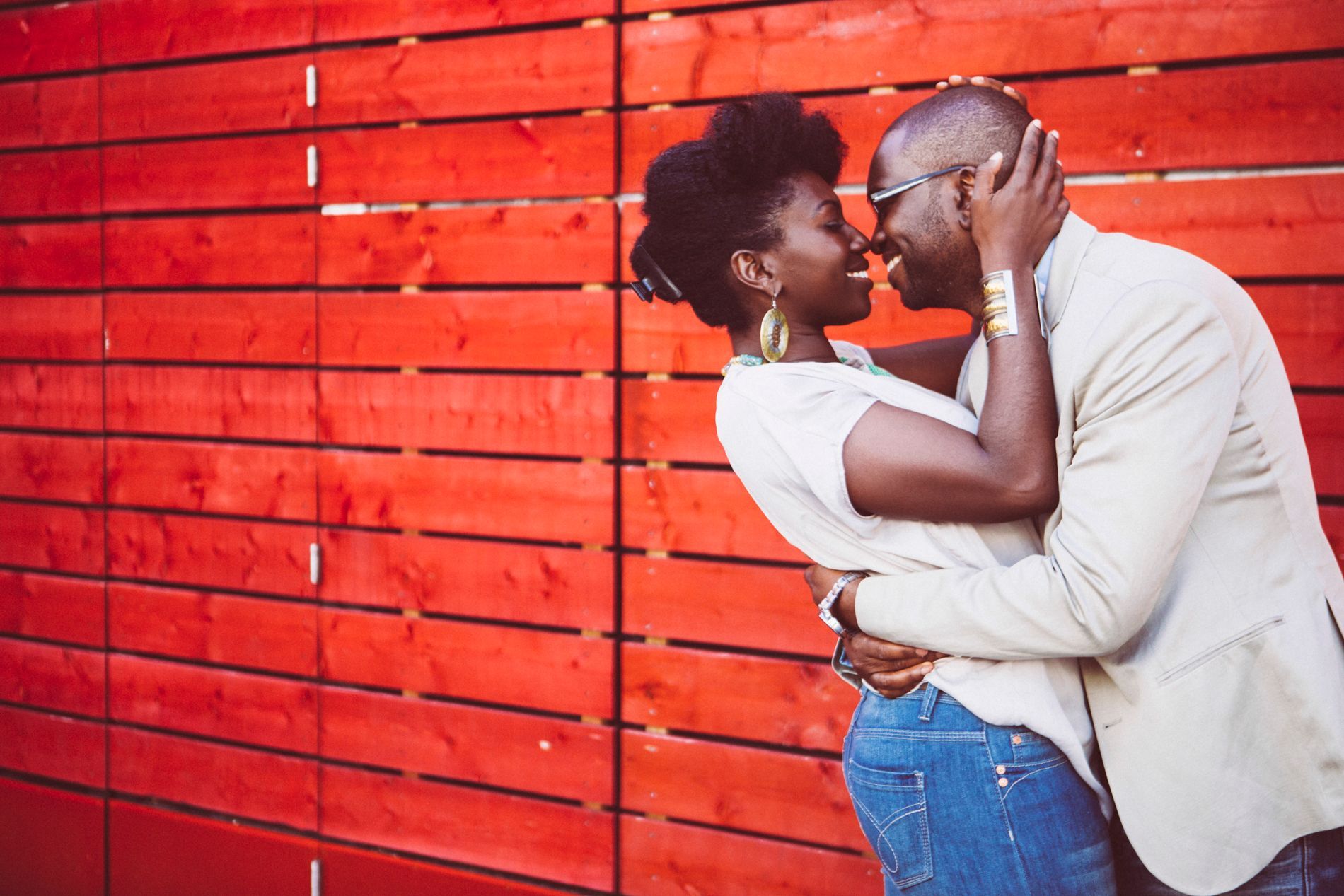 A man and a woman are kissing in front of a red wooden wall.
