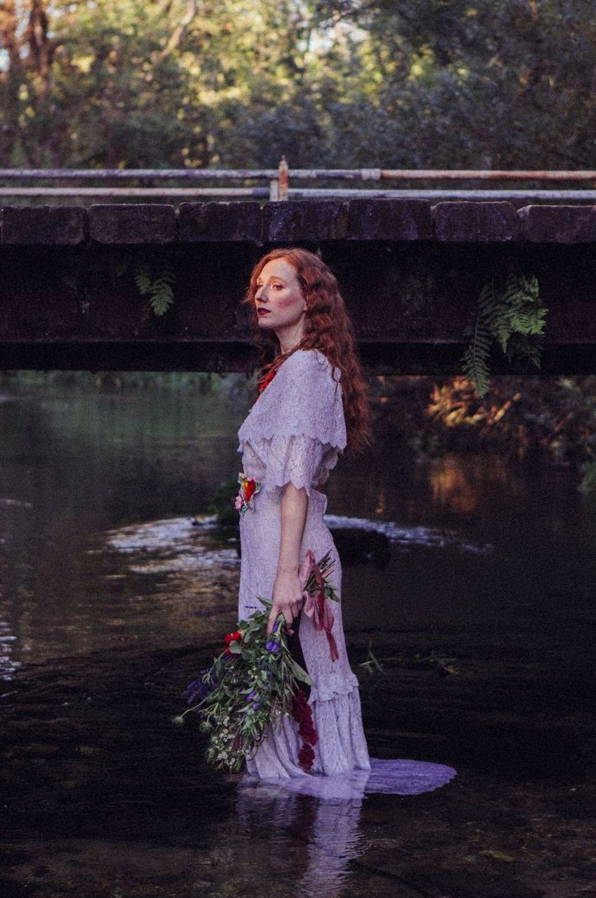 A woman in a white dress is standing in a river holding a bouquet of flowers.