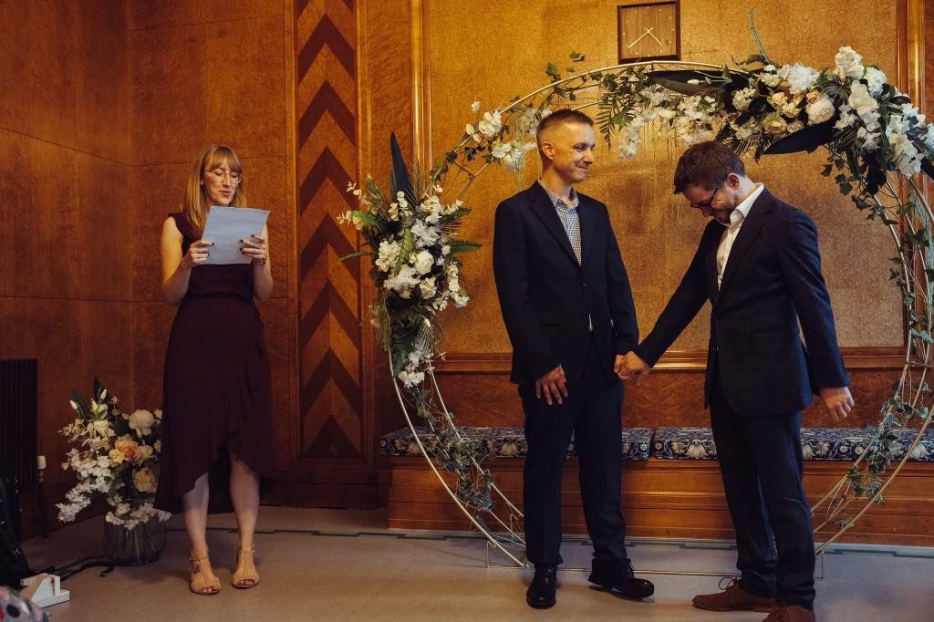 A woman is giving a speech at a wedding ceremony while two men hold hands.