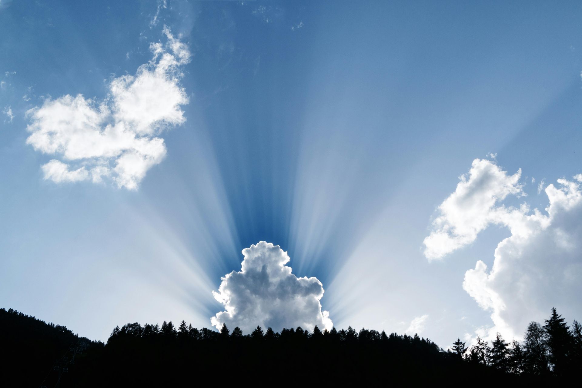 Sunlight bursts through clouds above a dark treeline, illuminating the blue sky.