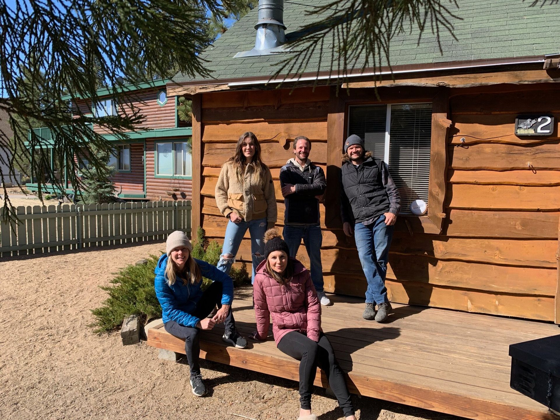 Five people pose on a wooden cabin's porch. Sunny day.
