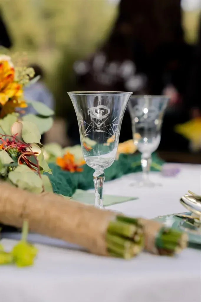 Two etched crystal wine glasses on a table set for dining, with a floral bouquet and wrapped stems.