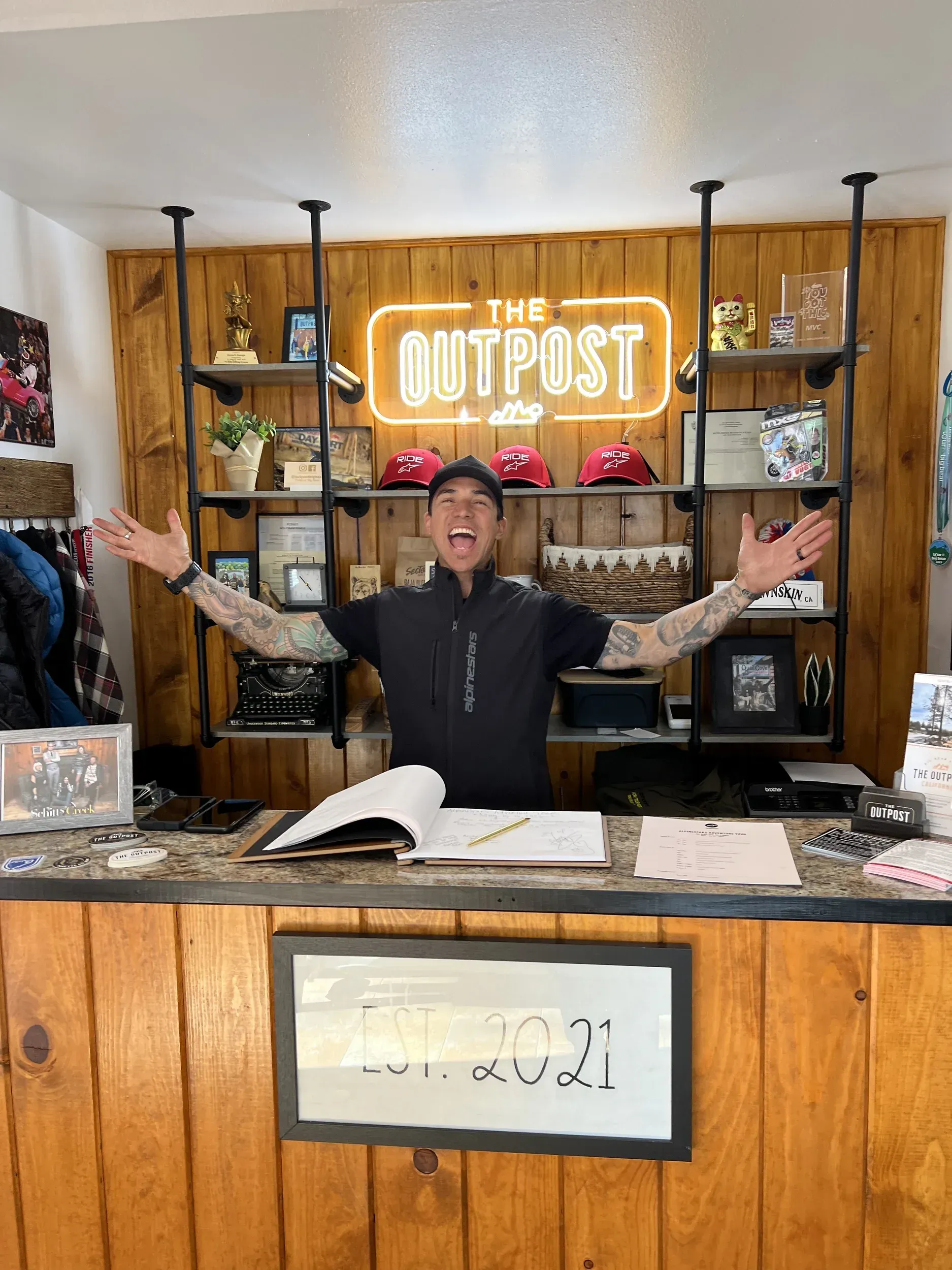 Man at The Outpost store counter with arms raised, smiling. Wooden shelves and sign