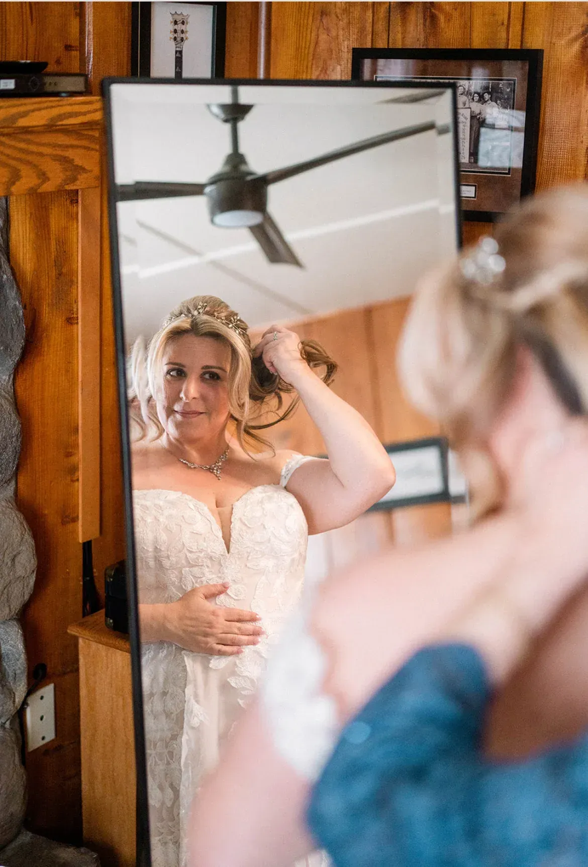 Bride in a white wedding dress adjusting her hair in a mirror, blue top in foreground, rustic wooden room.
