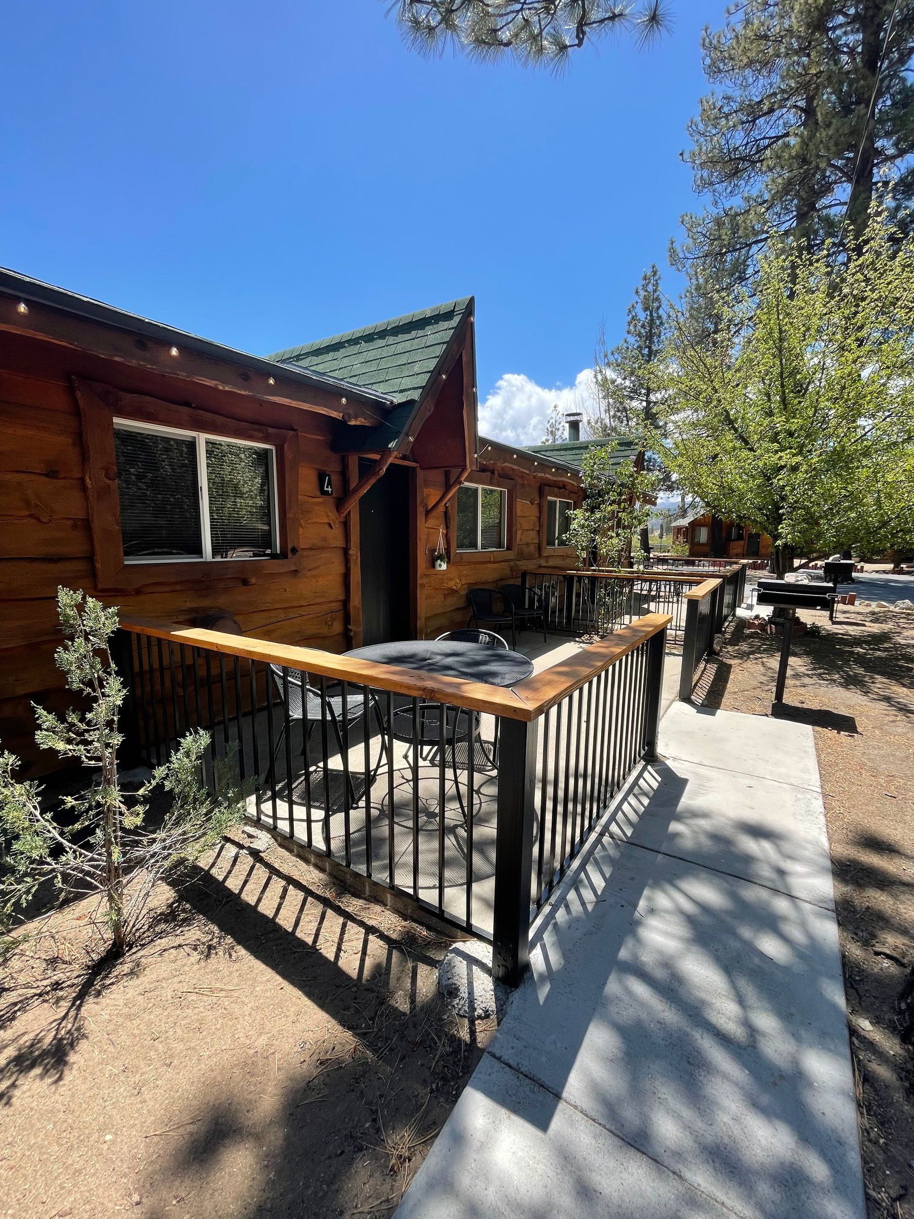 Log cabin with wooden deck and railings; trees in the background, blue sky.
